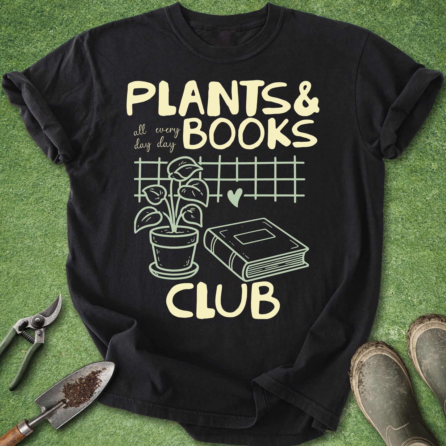 A black short-sleeve graphic T-shirt laid flat on green artificial grass, with gardening tools and boots around it. The shirt reads “PLANTS & BOOKS all every day CLUB” with a plant, book, heart, and grid illustration in pale yellow.