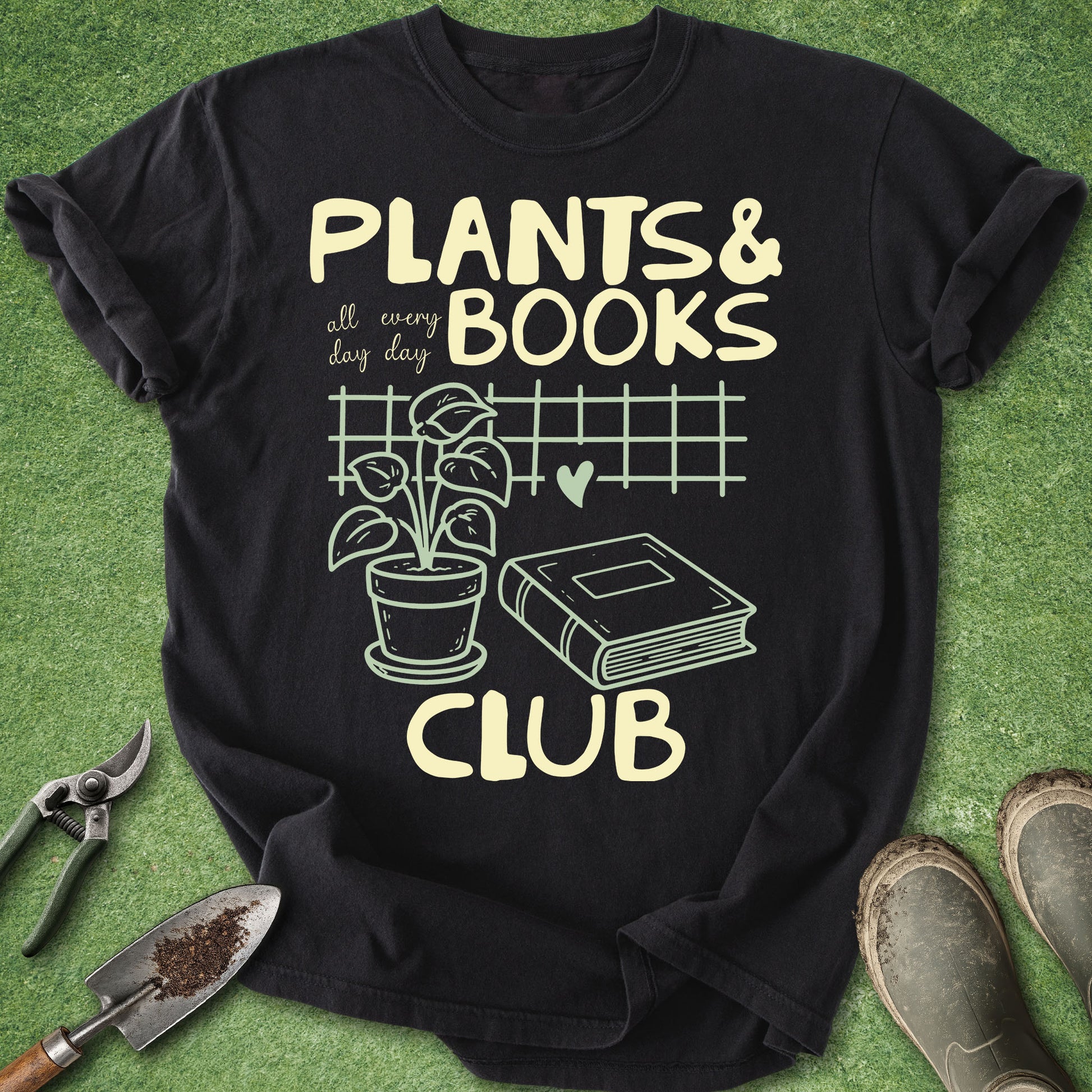 A black short-sleeve graphic T-shirt laid flat on green artificial grass, with gardening tools and boots around it. The shirt reads “PLANTS & BOOKS all every day CLUB” with a plant, book, heart, and grid illustration in pale yellow.