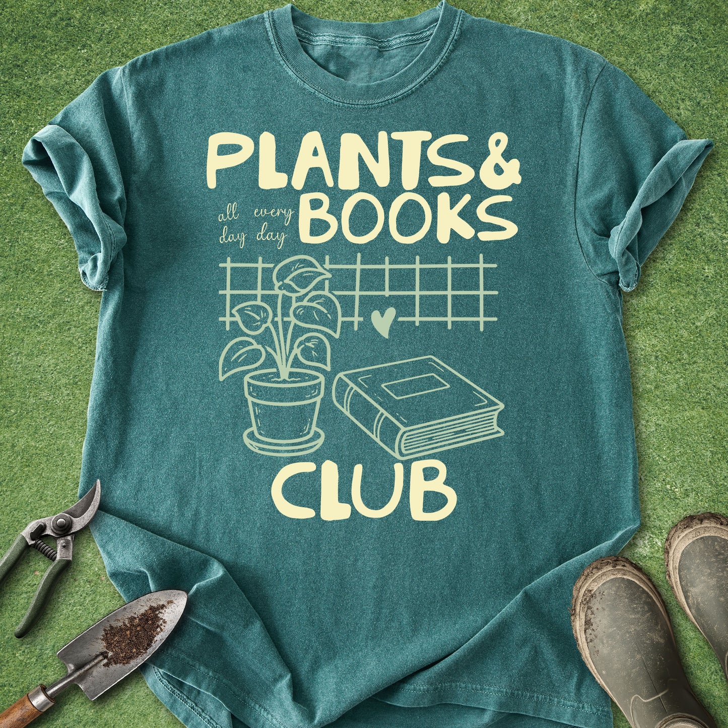 A teal short-sleeve graphic T-shirt laid flat on a green grass-like surface, with gardening tools and boots around it. The shirt reads “PLANTS & BOOKS all every day CLUB” in pale yellow text with a plant, pot, book, and heart illustration on the front.