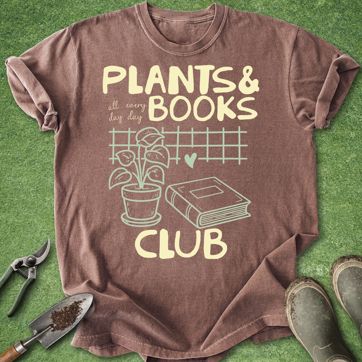 A brown short-sleeve graphic T-shirt laid flat on green grass-like background, with gardening tools and boots around it. The shirt reads 'PLANTS & BOOKS CLUB' with a plant, book, and grid illustration in pale yellow.