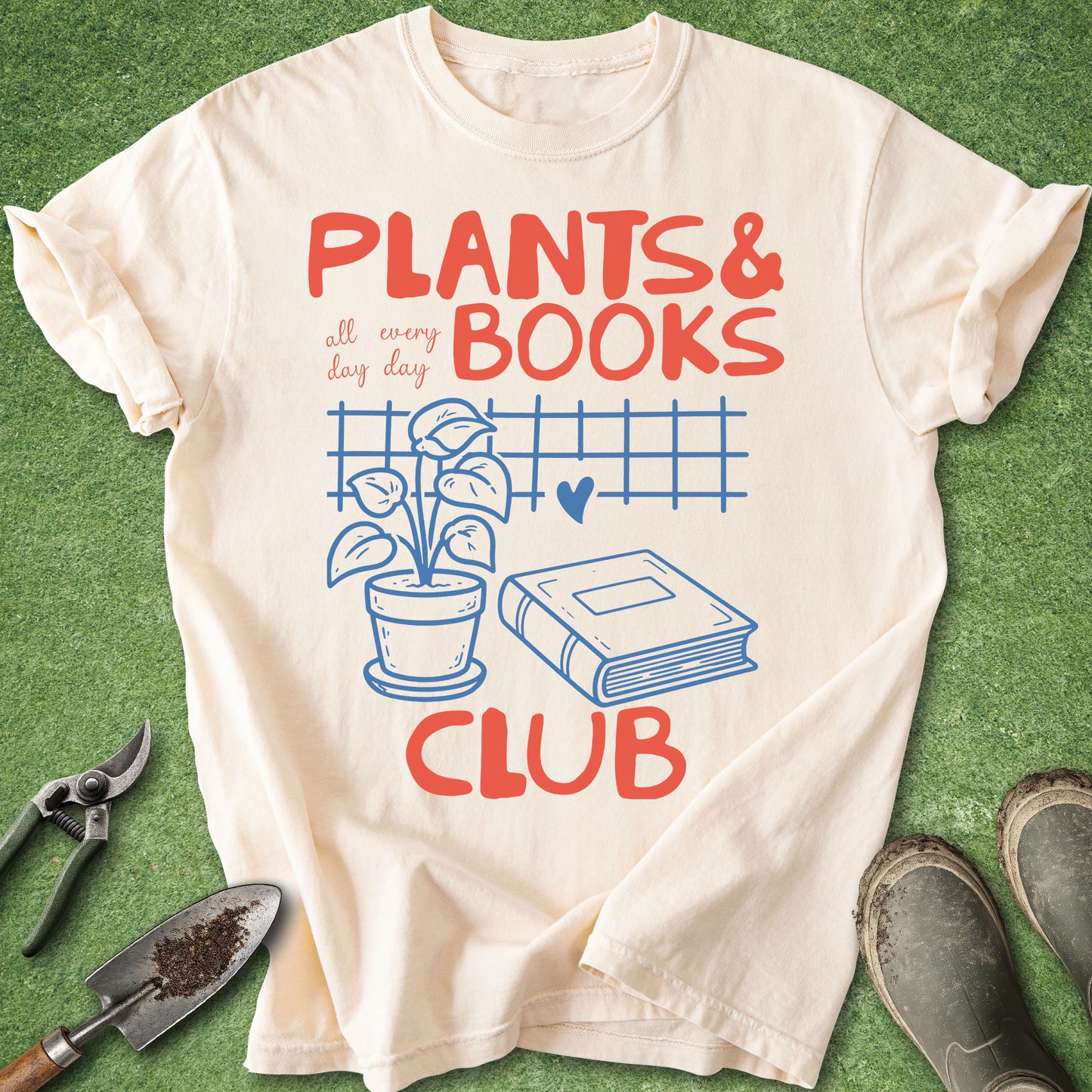 An off-white graphic t-shirt laid flat on green grass-like artificial turf, surrounded by gardening tools (shears, trowel) and muddy boots. The shirt features a red and blue graphic that reads 'PLANTS & BOOKS all every day CLUB' with an illustration of a potted plant and a book.