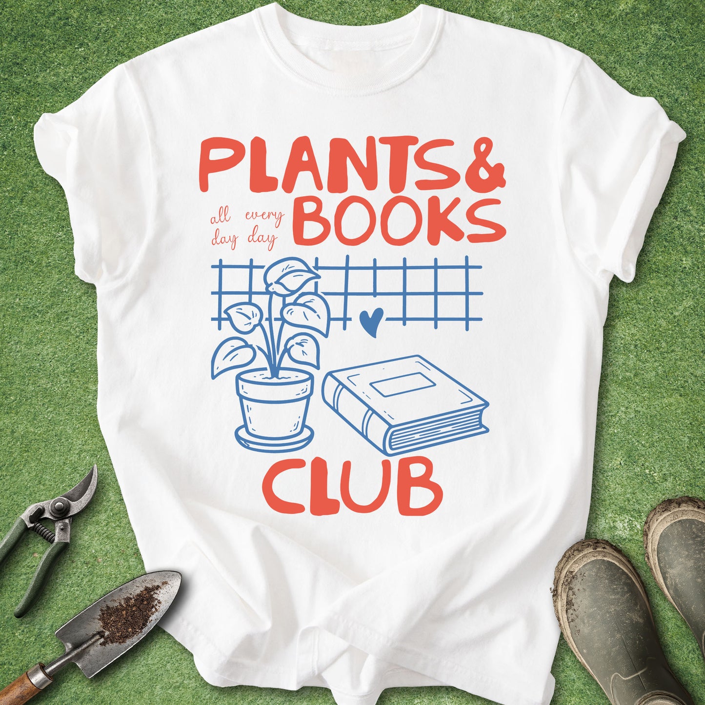 A white graphic T-shirt laid flat on green grass-like background, with red and blue text reading "PLANTS & BOOKS CLUB" and an illustration of a potted plant, book, and heart. Gardening tools and boots are placed around the shirt as styled props.