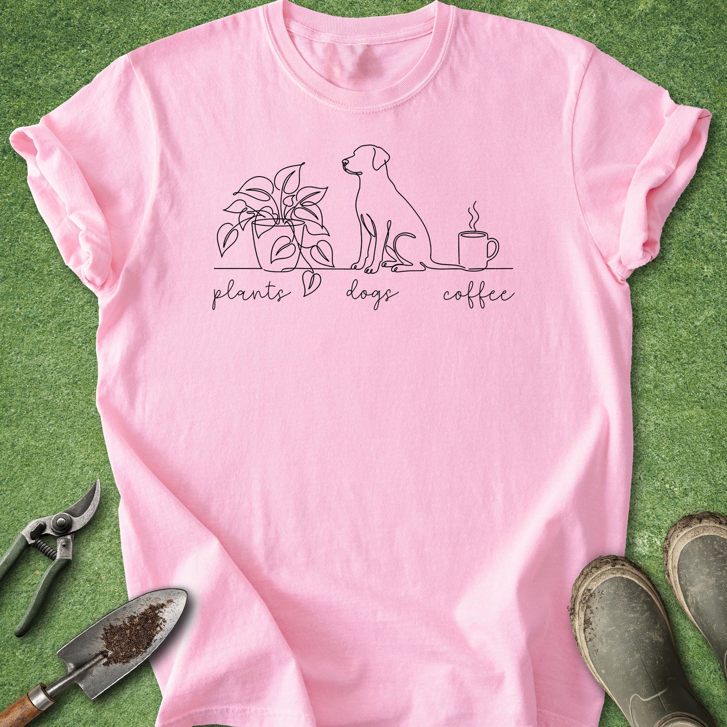 A pink short-sleeve t-shirt laid flat on green grass-like artificial turf, featuring a black line art graphic of a plant, a dog, and a coffee mug with the words 'plants dogs coffee' underneath. Gardening tools and muddy boots are placed around the shirt as styling props.