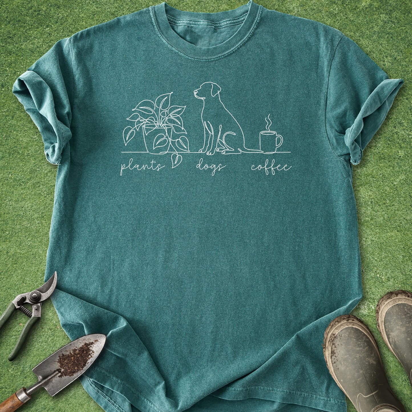 A teal short-sleeve t-shirt with a white line art graphic of a dog, a plant, and a coffee mug with the words 'plants dogs coffee' printed on the front. The shirt is laid flat on green grass with gardening tools (trowel, pruning shears) and boots as props.