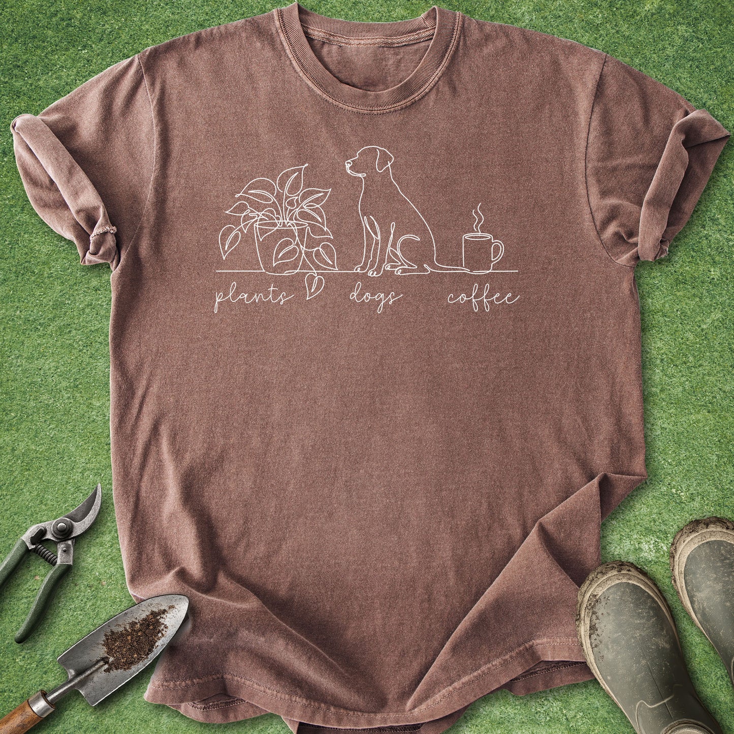 A brown short-sleeve t-shirt with a white line art graphic of a dog, a plant, and a coffee mug, with the text 'plants + dogs + coffee' printed below. The shirt is laid flat on a green grass-like surface with gardening tools and boots around it.
