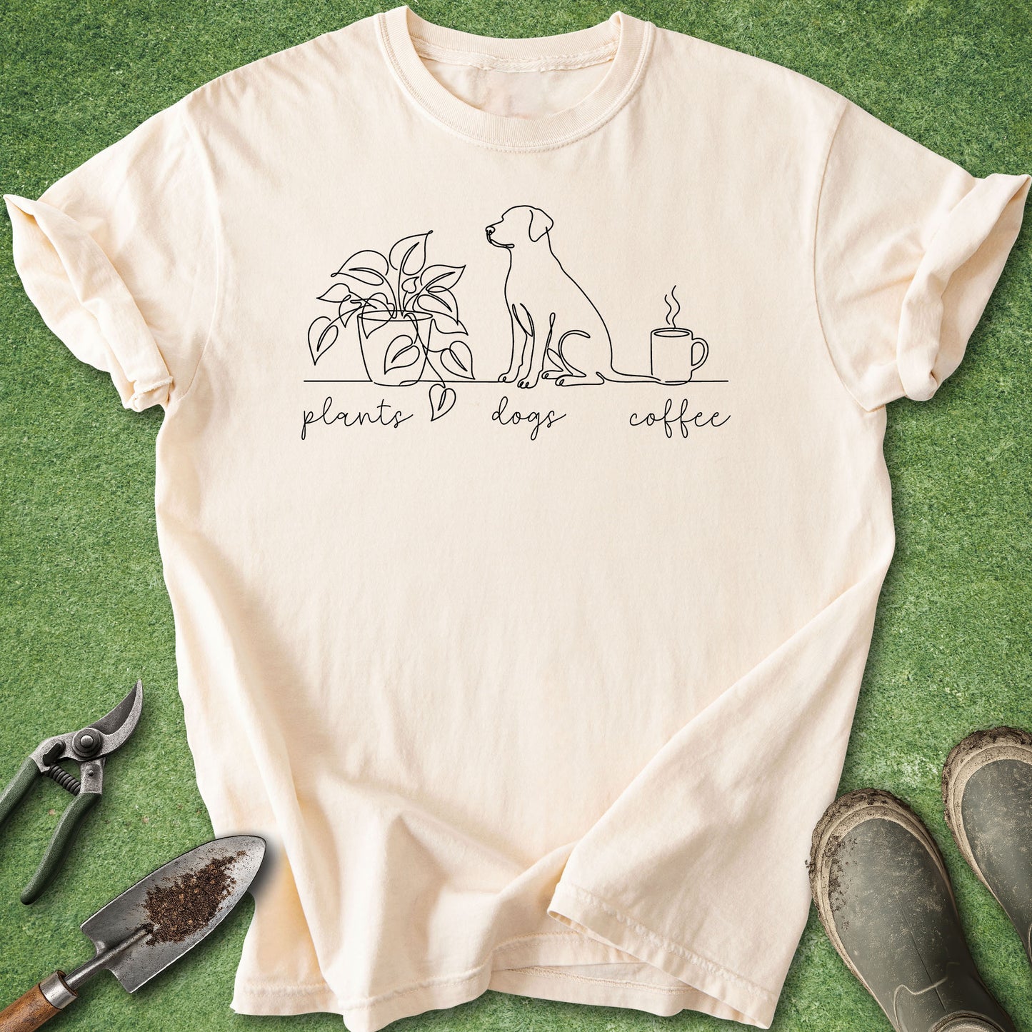 A cream-colored short-sleeve t-shirt with a black line art graphic of a dog, a plant, and a coffee mug, with the words 'plants', 'dogs', and 'coffee' written below. The shirt is laid flat on a green grass-like surface with gardening tools (trowel, pruning shears) and muddy boots nearby.