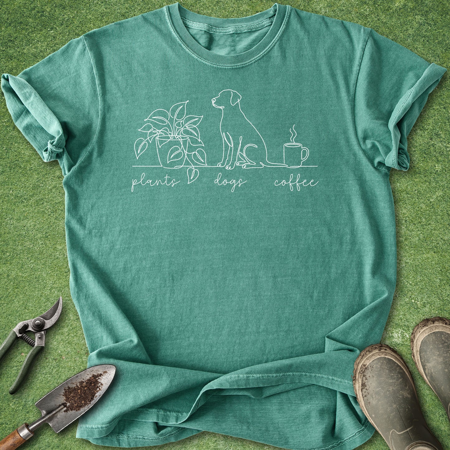 A sage green short-sleeve t-shirt with a white line art graphic of a plant, a dog, and a coffee cup, with the words 'plants > dogs > coffee' printed below. The t-shirt is laid flat on green grass-like artificial turf with gardening tools (trowel, pruning shears) and muddy boots as props.