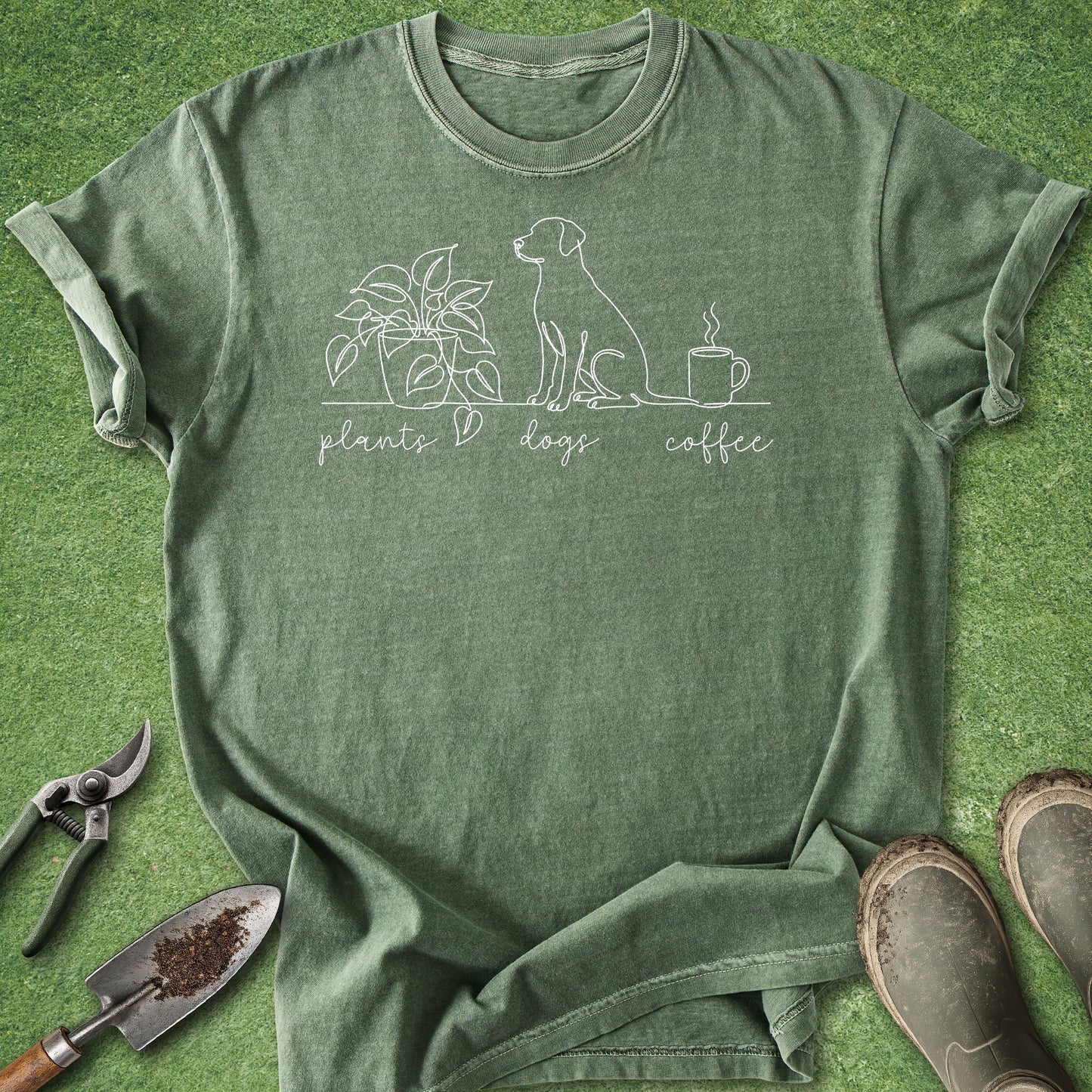 An olive green heathered t-shirt with a white line art graphic of a plant, a dog, and a coffee cup with the words 'plants dogs coffee' printed on the front. The shirt is laid flat on a green grass-like surface, styled with gardening tools (trowel, pruning shears) and muddy boots.