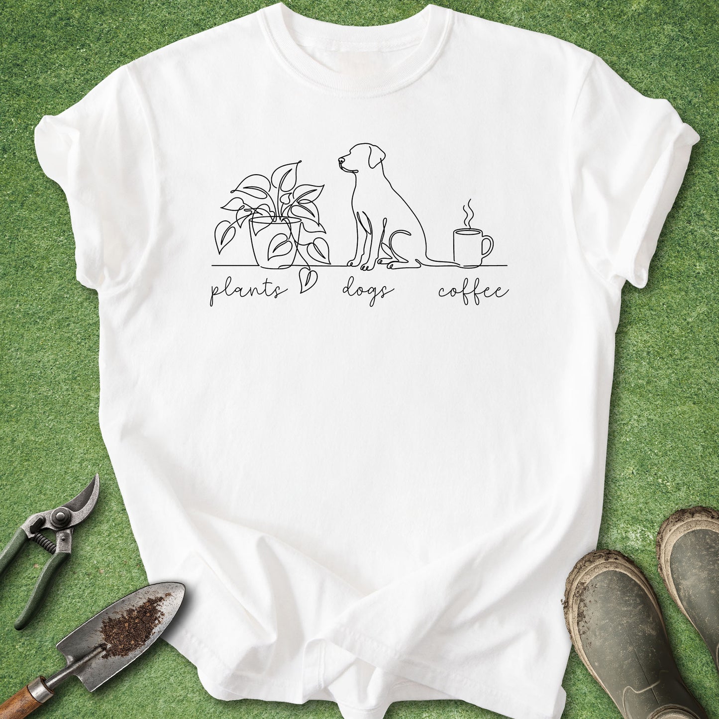 A white short-sleeve graphic T-shirt laid flat on green grass-like background, with a simple black line drawing of a plant, a dog, and a coffee mug above the words 'plants dogs coffee.' Gardening tools and muddy boots are placed around the shirt as styling props.