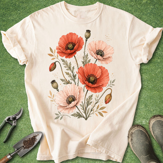An off-white short-sleeve t-shirt with a watercolor-style poppy floral print, laid flat on green grass with gardening props like pruning shears, a trowel, and boots around it.