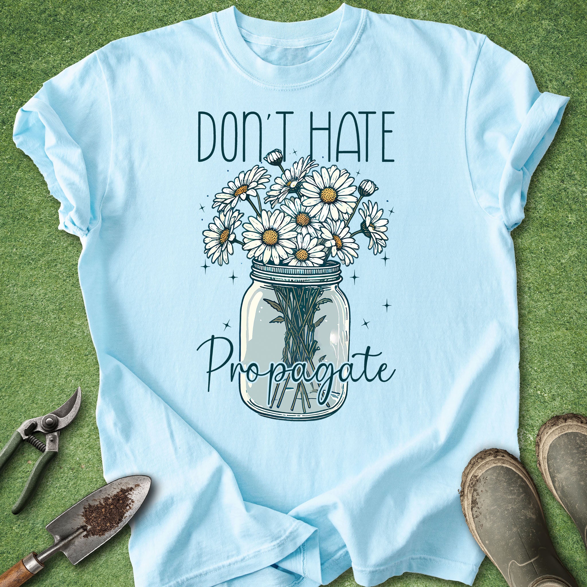 Light blue t-shirt with a graphic of flowers in a jar and text 'Don't Hate Propagate' on a green grass background.