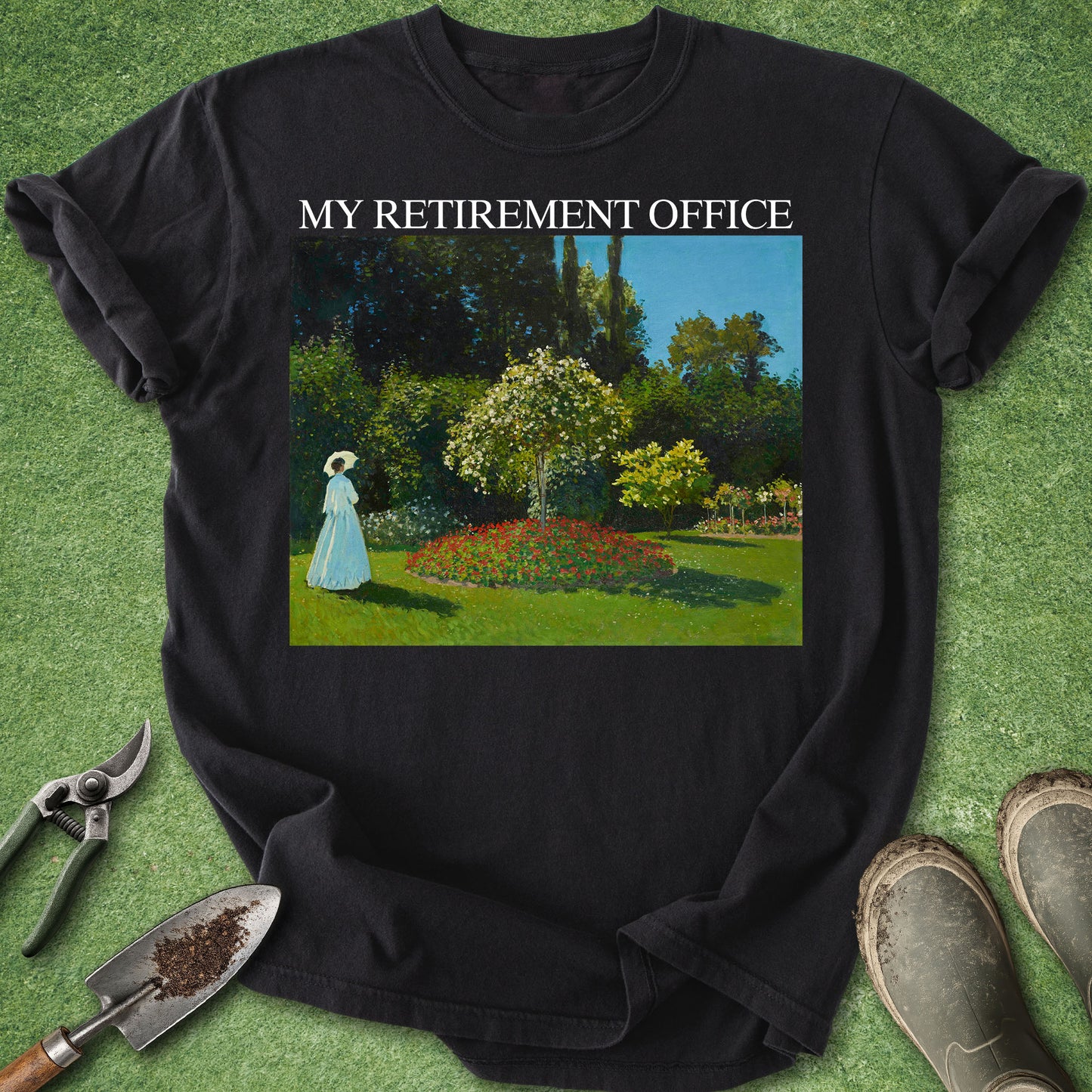 A black short-sleeve graphic T-shirt laid flat on a green grass-like surface, with gardening tools and boots around it. The shirt has white text reading 'MY RETIREMENT OFFICE' above a rectangular garden scene print showing a woman in a blue dress standing in a flower bed.