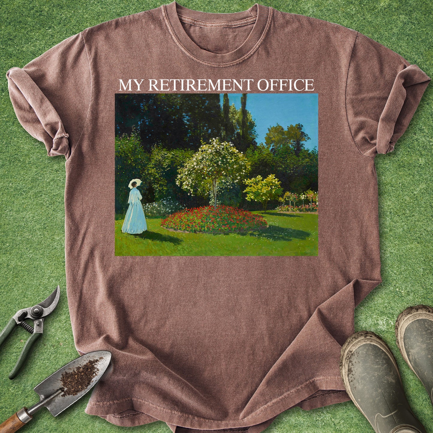 A brown short-sleeve t-shirt laid flat on a green grass-like surface, featuring a square graphic print of a garden scene with the text 'MY RETIREMENT OFFICE' above it. Gardening tools and boots are placed around the shirt as styling props.