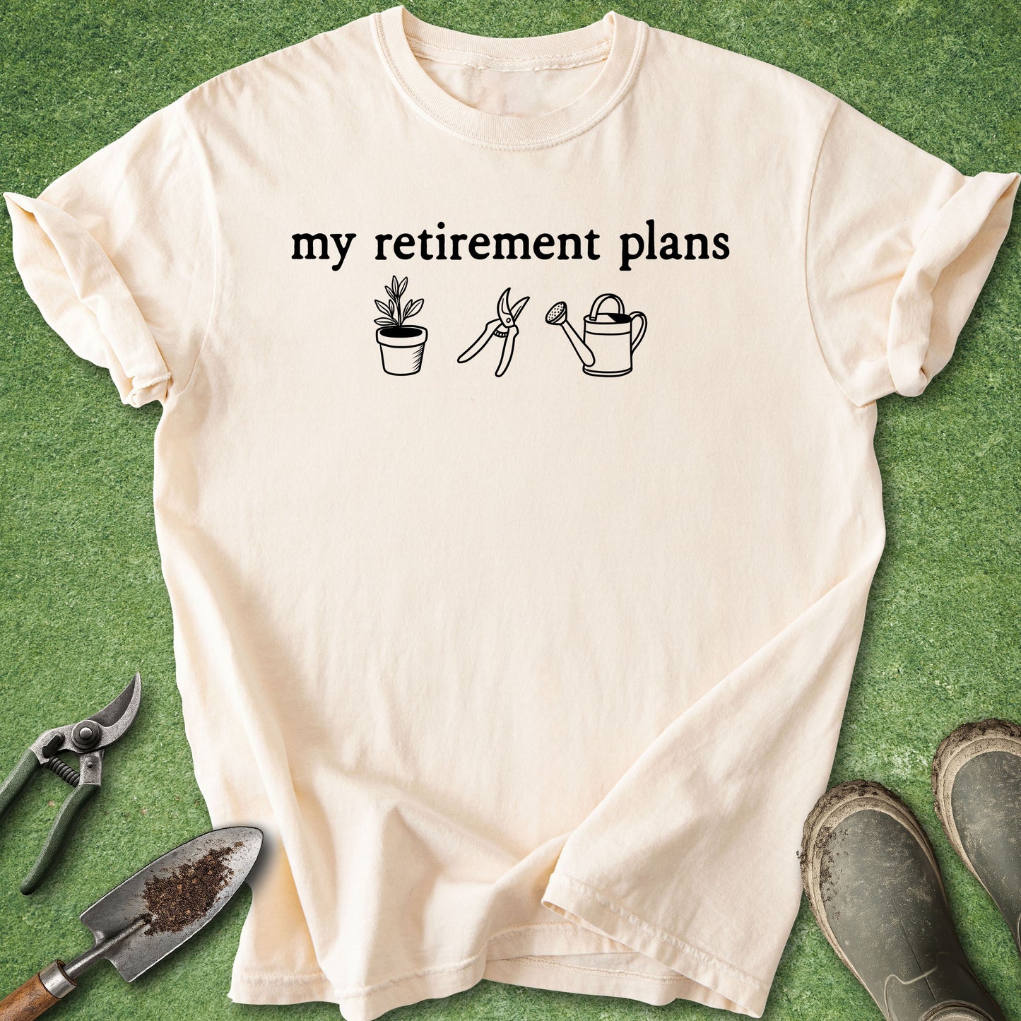 A cream-colored short-sleeve t-shirt with the black text 'my retirement plans' and simple black line drawings of a potted plant, pruning shears, and watering can, laid flat on a green grass-like surface with gardening tools and muddy boots nearby.