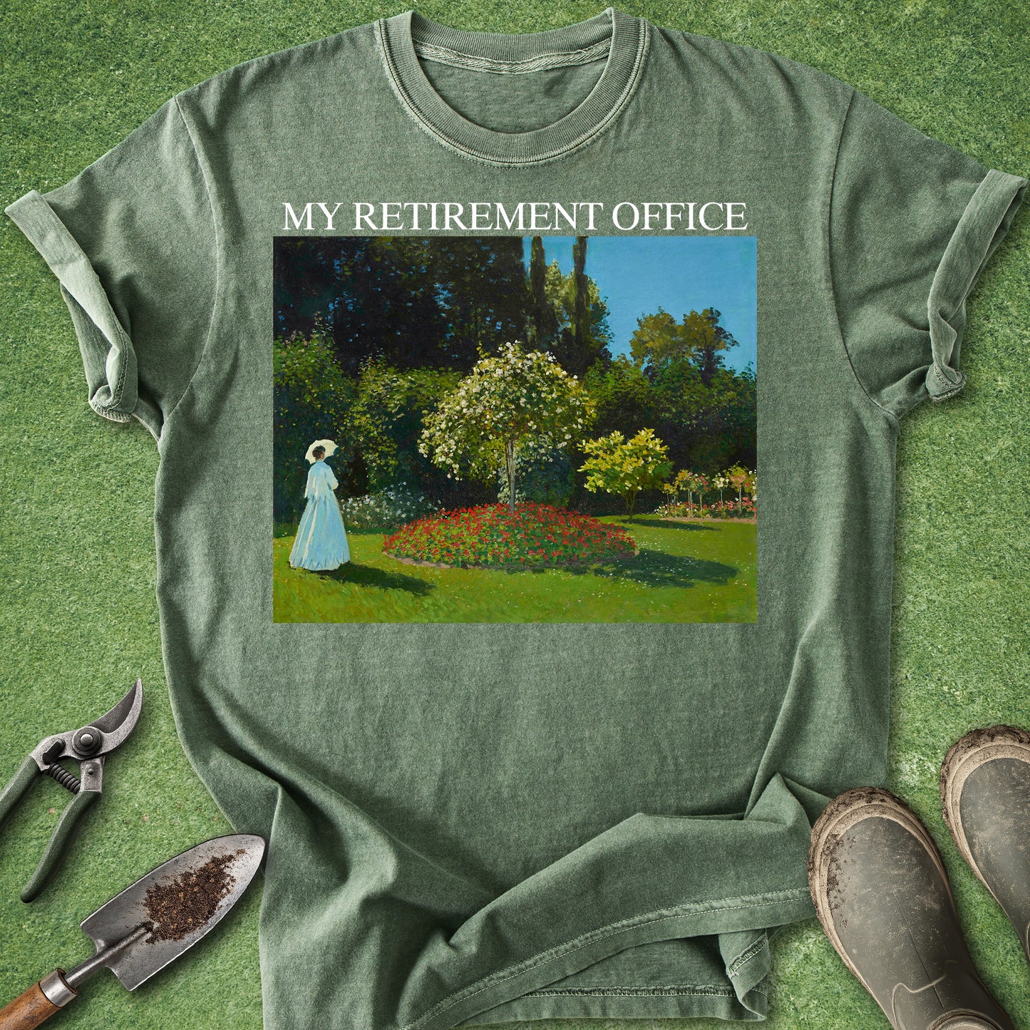 An olive green graphic t-shirt laid flat on a green grass-like surface, surrounded by gardening props including pruning shears, a trowel, and boots. The t-shirt features a square graphic print of a garden scene with the text 'MY RETIREMENT OFFICE' above it.