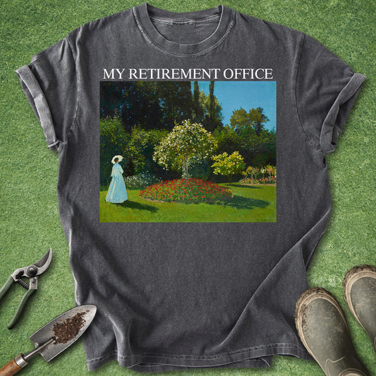 A dark gray graphic t-shirt laid flat on a green grass-like surface, with gardening tools and boots around it. The shirt has the text 'MY RETIREMENT OFFICE' above a colorful garden scene print.