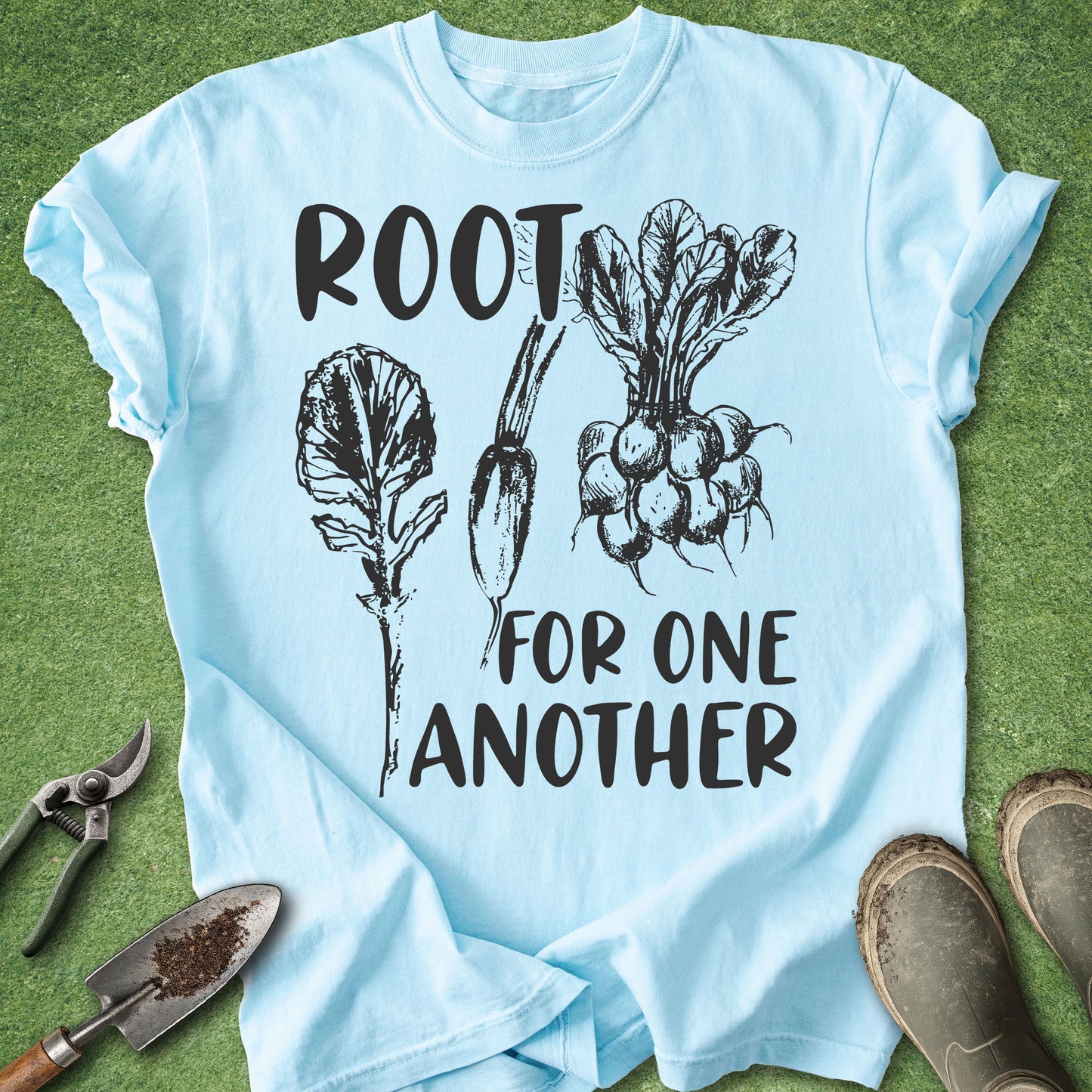 A light blue graphic t-shirt laid flat on green grass, featuring black text that reads 'ROOT FOR ONE ANOTHER' with illustrations of leafy greens and radishes. Gardening tools and muddy boots are placed around the shirt as props.
