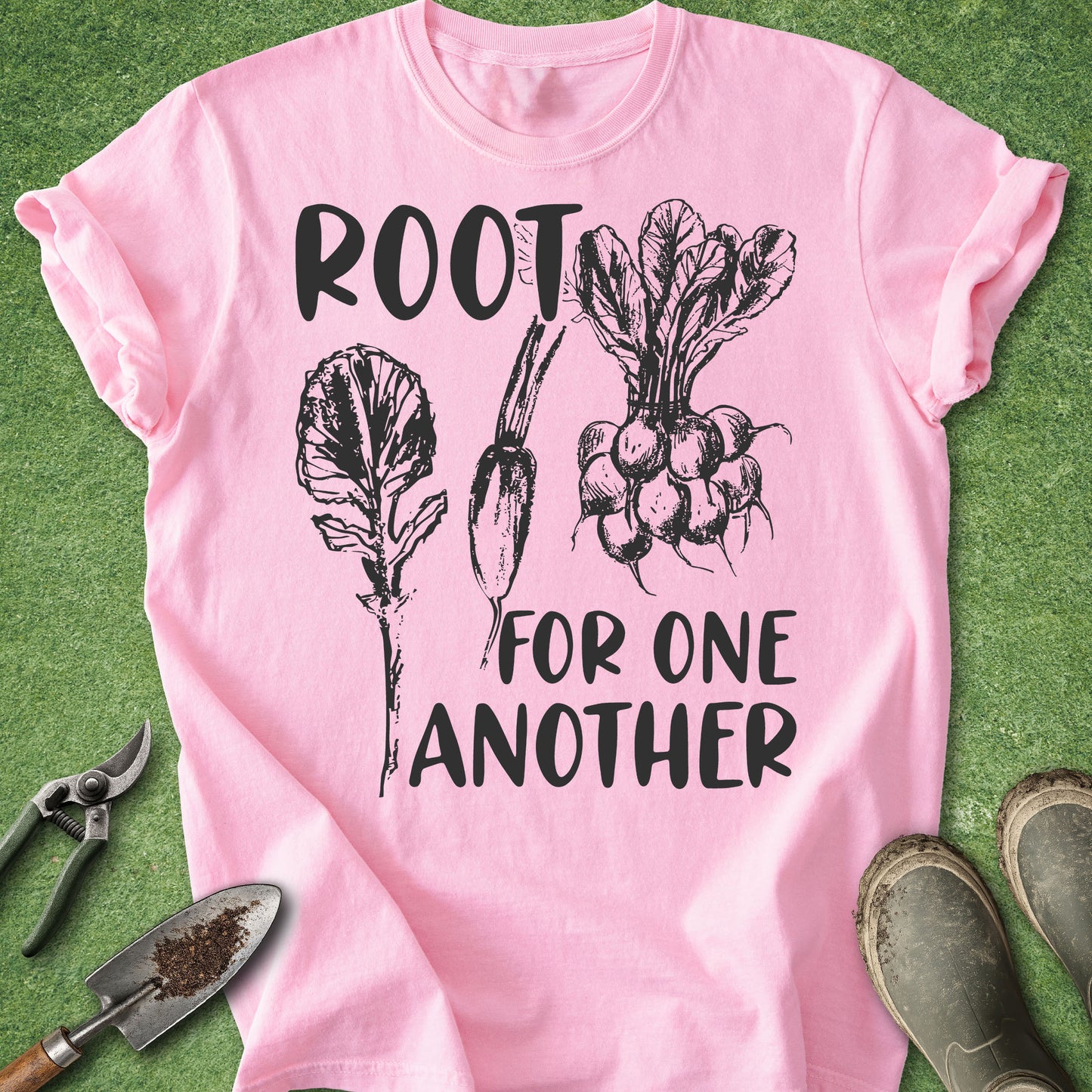 A pink short-sleeve T-shirt laid flat on green grass-like background, with black text reading 'ROOT FOR ONE ANOTHER' and black line drawings of leafy greens, a carrot, and radishes. Gardening tools and boots are placed around the shirt as styling props.