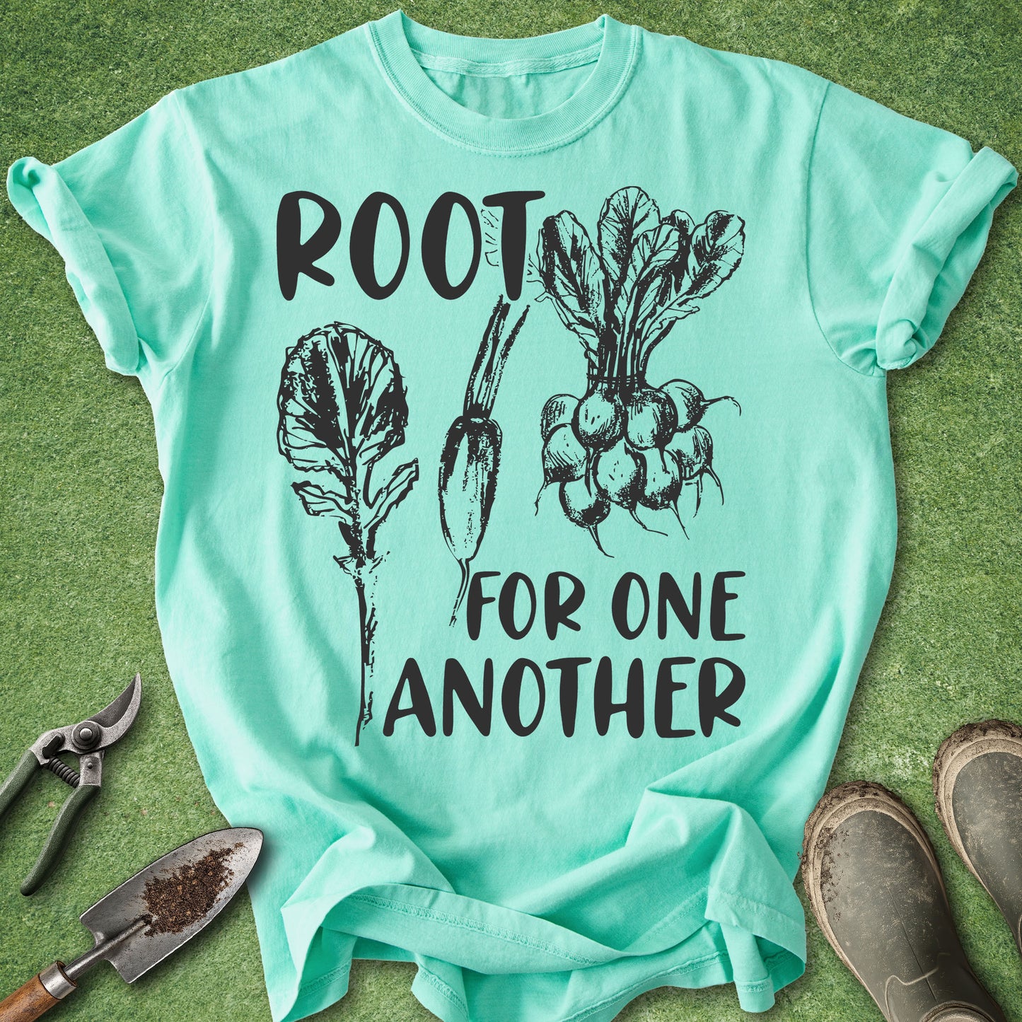 A mint green graphic t-shirt laid flat on a green grass-like surface, featuring black text that reads 'ROOT FOR ONE ANOTHER' with illustrations of vegetables. Gardening props including pruning shears, a trowel, and boots are arranged around the shirt.