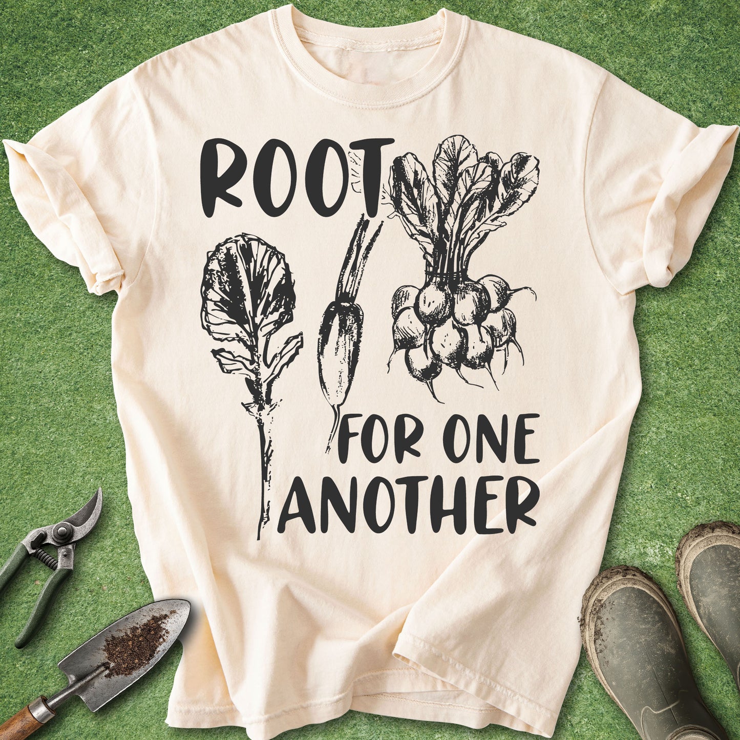 A cream-colored graphic t-shirt laid flat on a green grass-like surface, featuring black text that reads 'ROOT FOR ONE ANOTHER' with illustrations of leafy greens and radishes. Gardening tools and muddy boots are placed around the shirt as styling props.