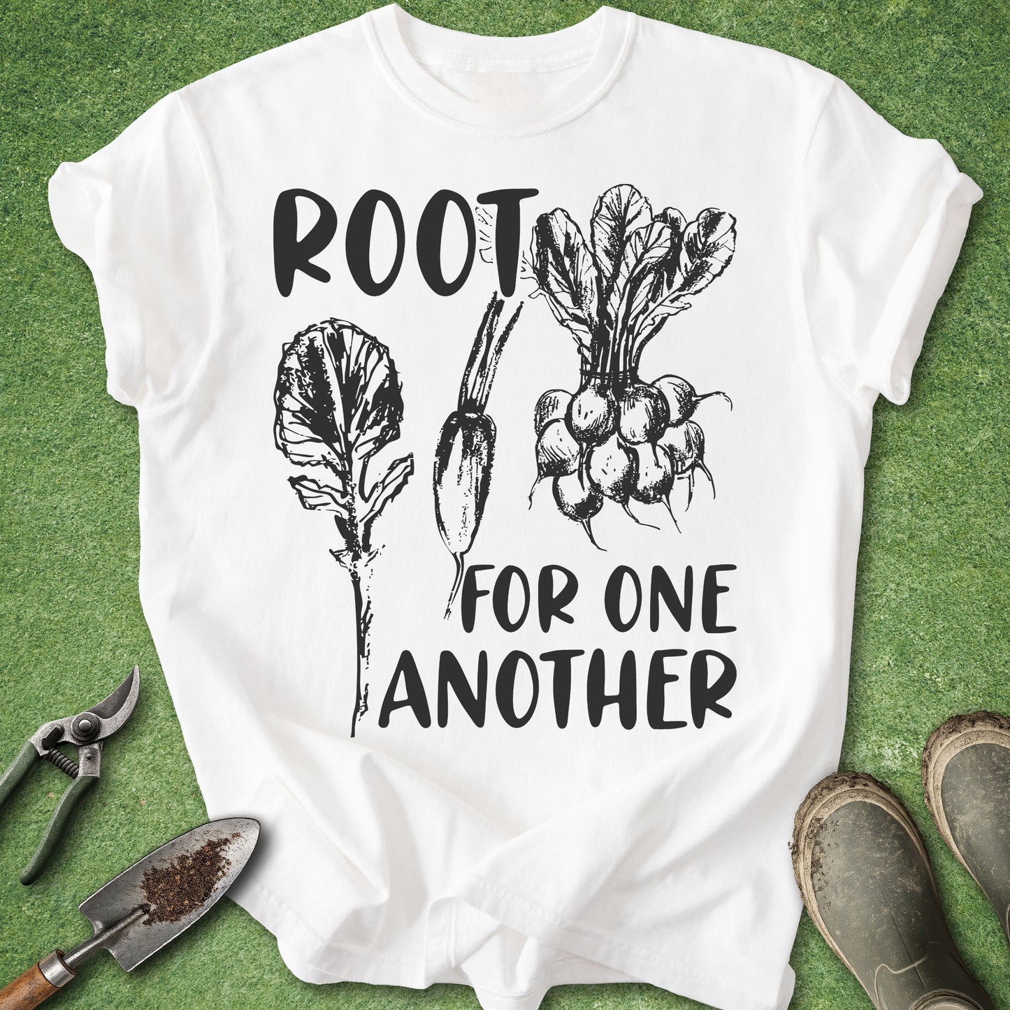 A white short-sleeve graphic T-shirt laid flat on green grass, with black text reading 'ROOT FOR ONE ANOTHER' and black line drawings of leafy greens and radishes. Gardening tools and muddy boots are partially visible around the shirt.