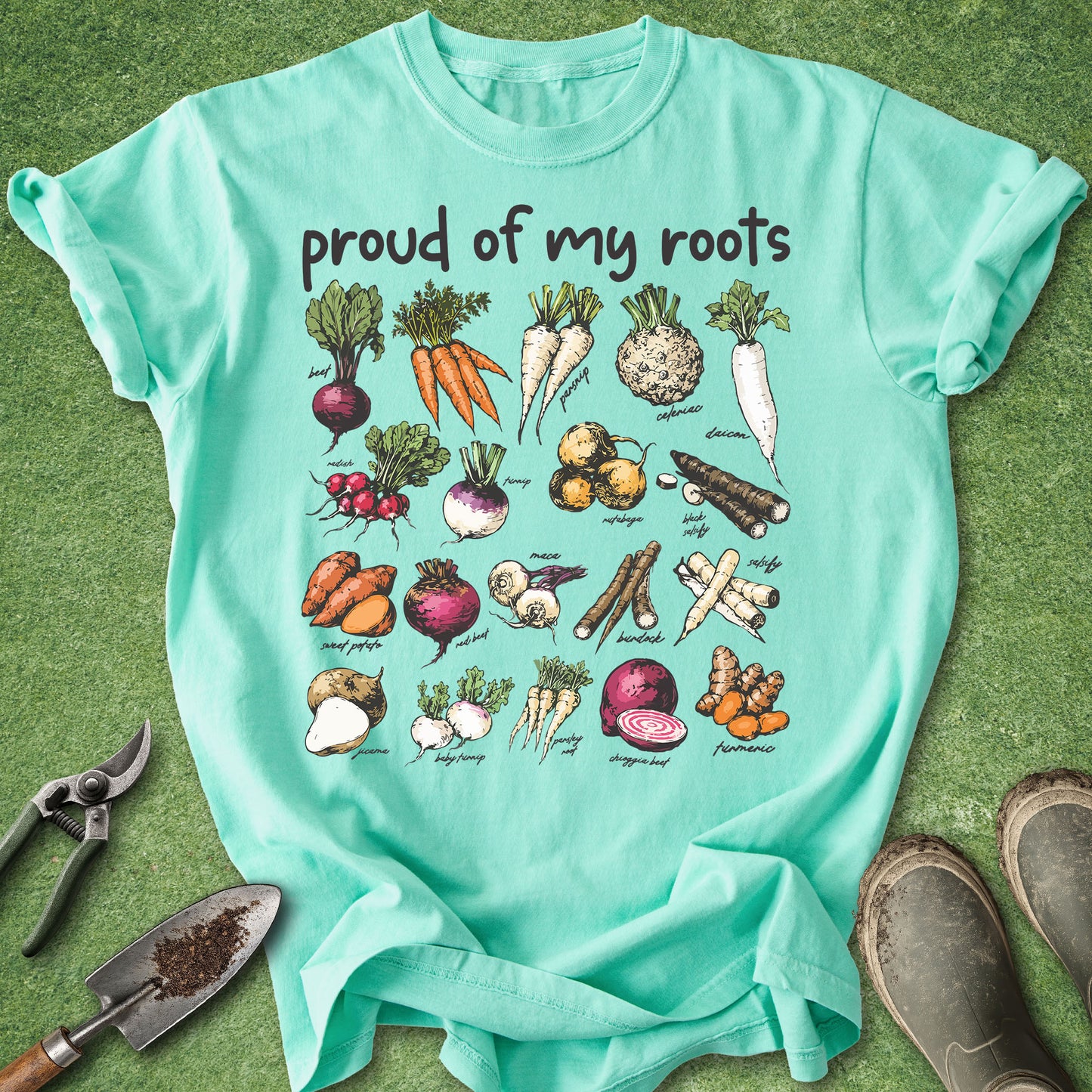 Mint green t-shirt with vegetable illustrations and 'proud of my roots' text on a grass background.