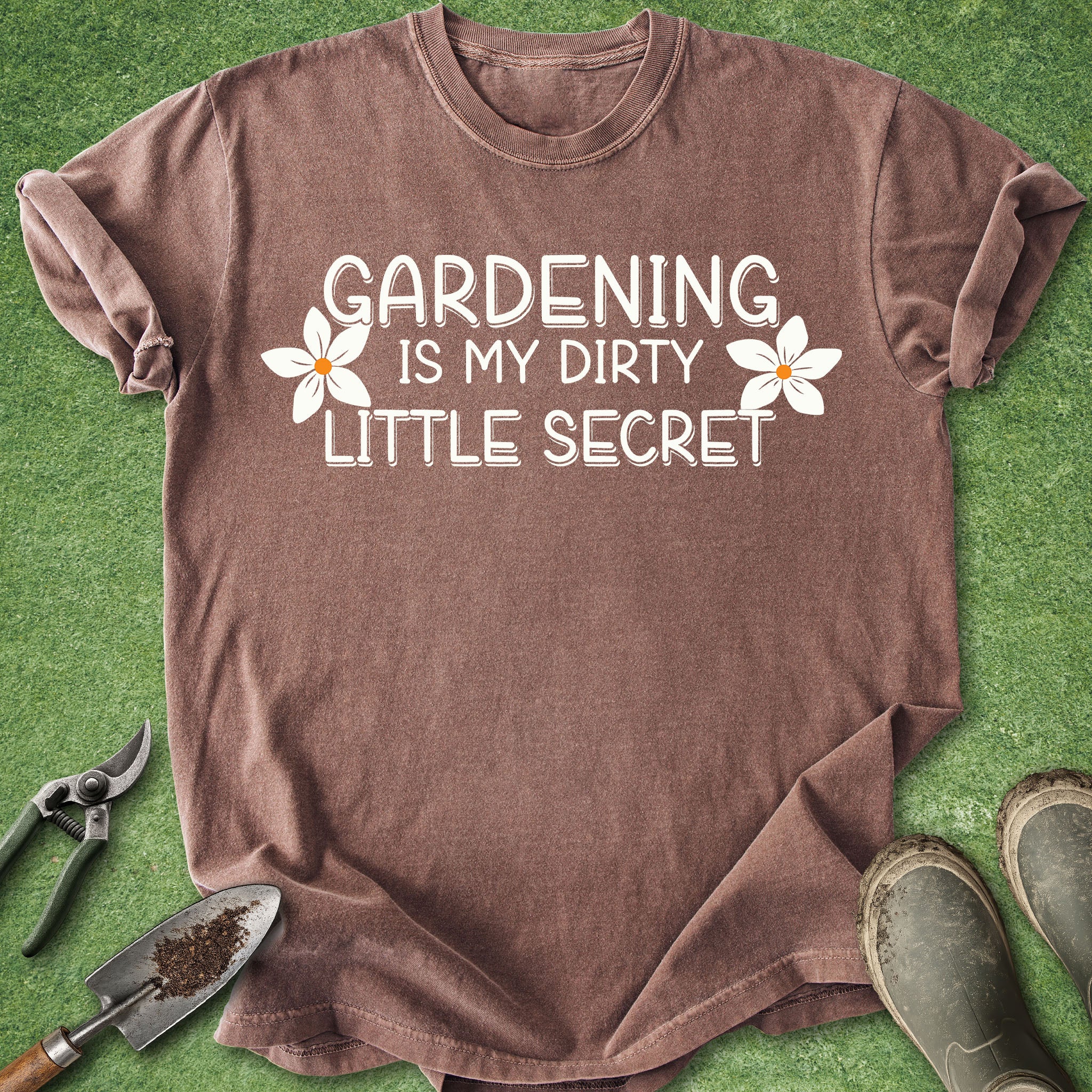 Brown t-shirt with 'Gardening is my dirty little secret' text on a grass background with gardening tools and boots.