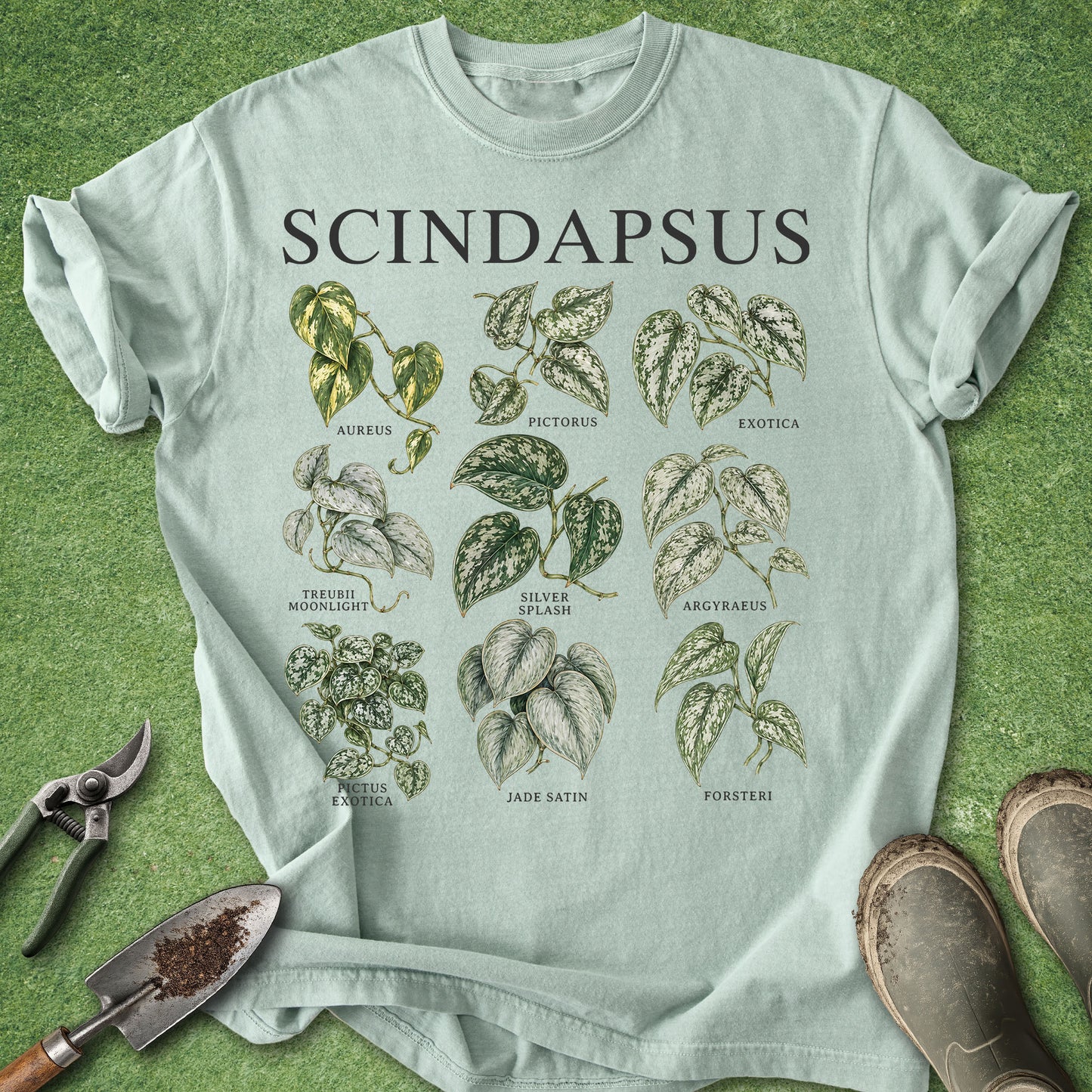 A light sage green t-shirt featuring a botanical graphic of various Scindapsus plant varieties with the word 'SCINDAPSUS' printed at the top. The shirt is styled in a flat-lay on a green grass-like surface, accompanied by gardening tools (shears, trowel) and a pair of boots.