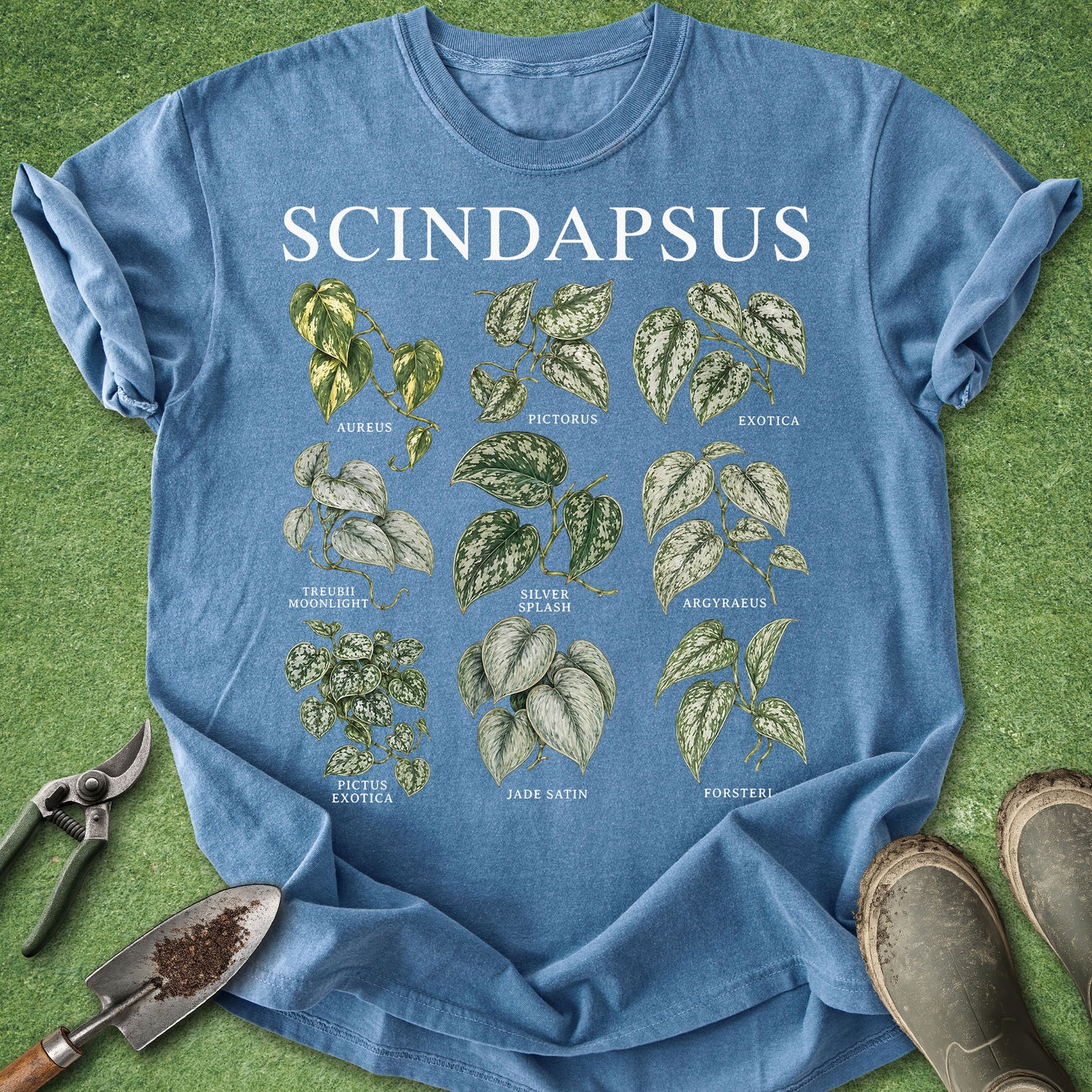 A blue short-sleeve graphic T-shirt laid flat on green grass, with gardening tools and boots around it. The shirt has large white text reading 'SCINDAPSUS' and a central botanical leaf chart in green, beige, and white.
