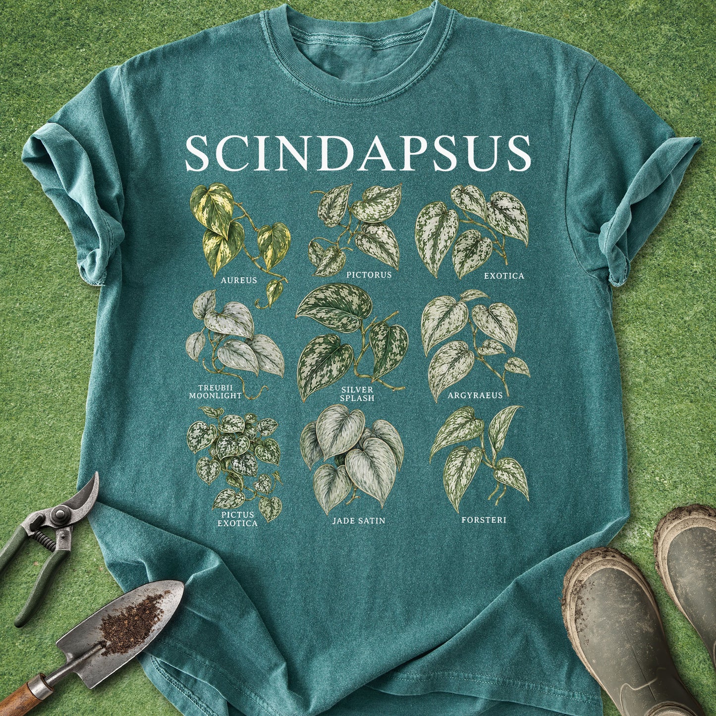 A teal short-sleeve graphic T-shirt laid flat on green grass, featuring the word 'SCINDAPSUS' in large white letters above illustrated plant leaves and small labels. Gardening props including pruning shears, a small hand trowel with soil, and boots are visible around the shirt.