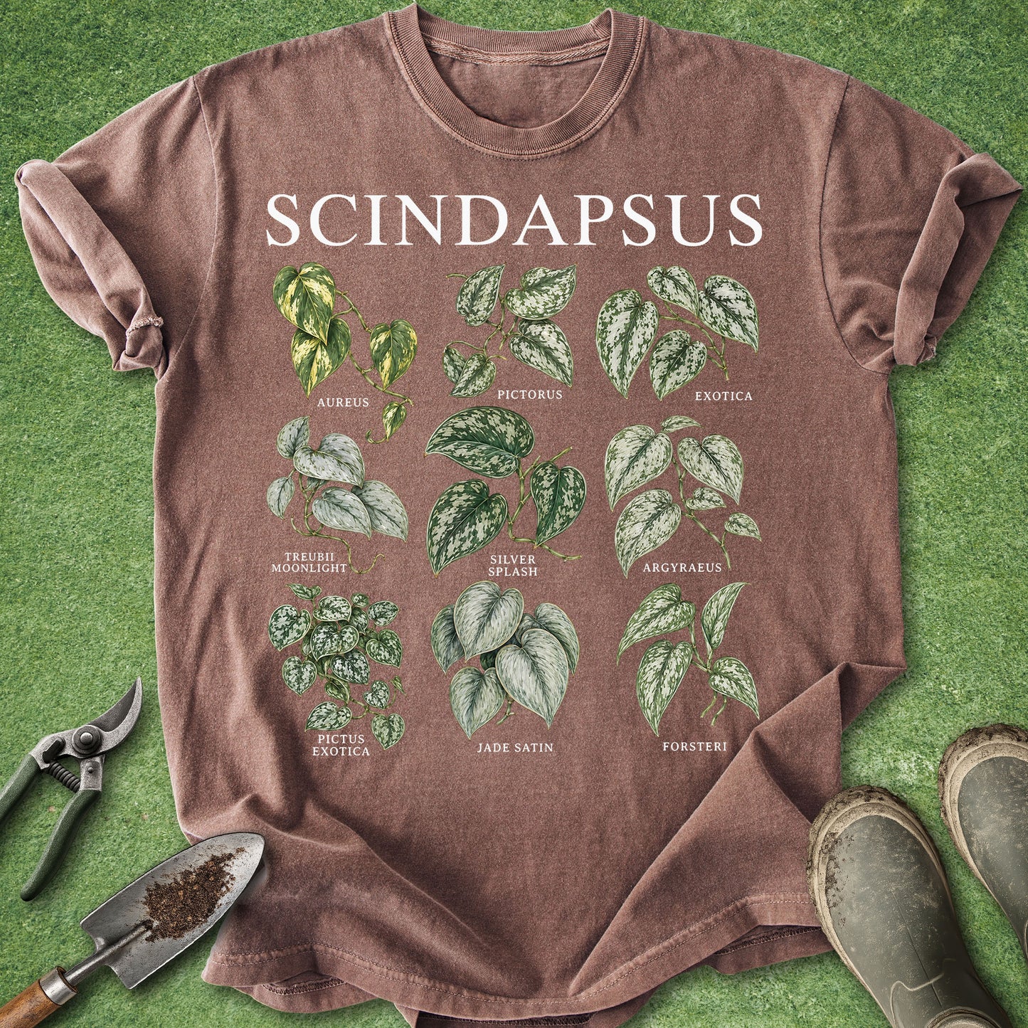 A brown short-sleeve graphic T-shirt laid flat on green grass, featuring the word 'SCINDAPSUS' and illustrated leafy plant labels. Gardening tools and boots are partially visible around the shirt.