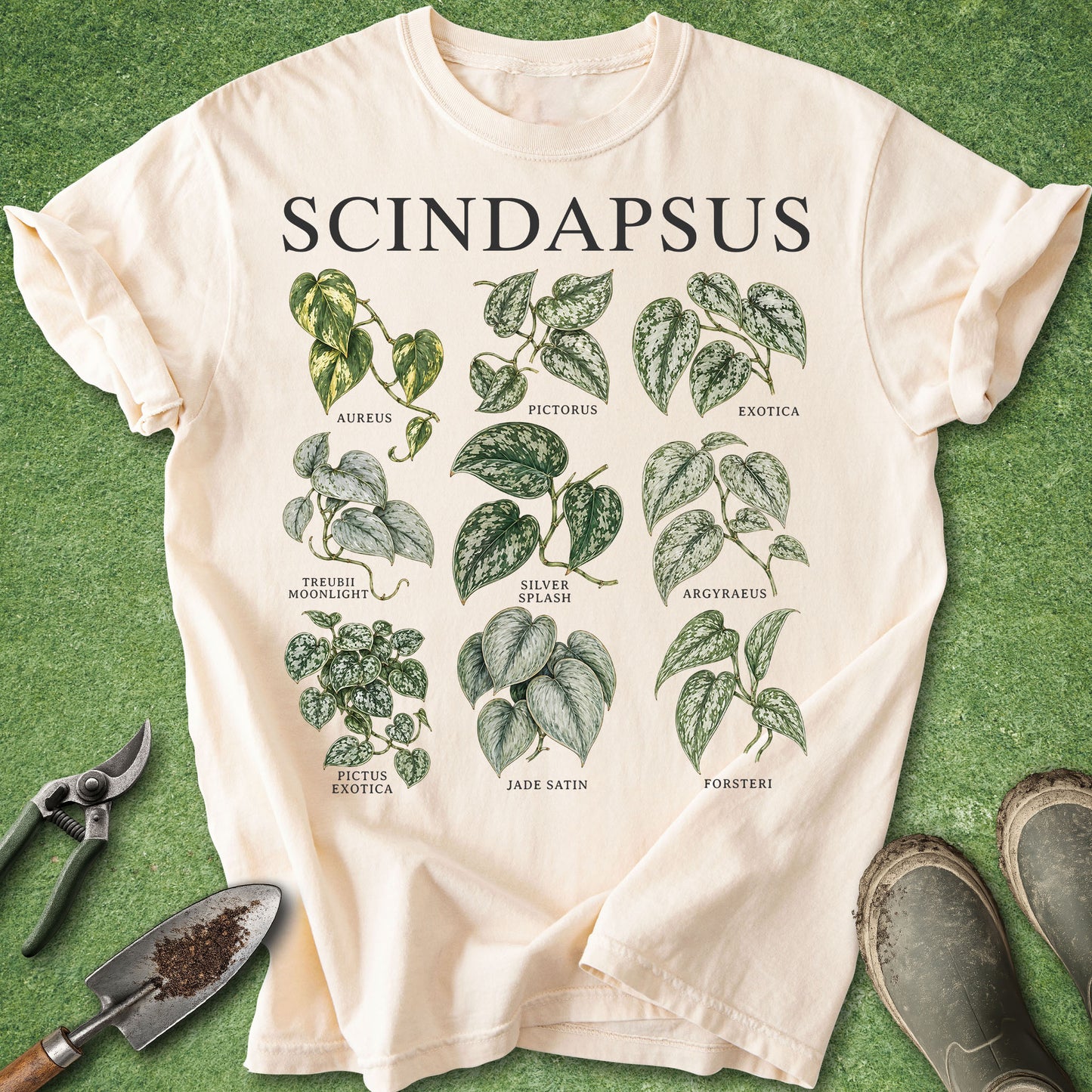 An off-white short-sleeve T-shirt laid flat on green grass-like ground, printed with the word 'SCINDAPSUS' and illustrated green leafy plant graphics with labels. Gardening props including pruning shears, a small trowel, and boots are placed around the shirt.