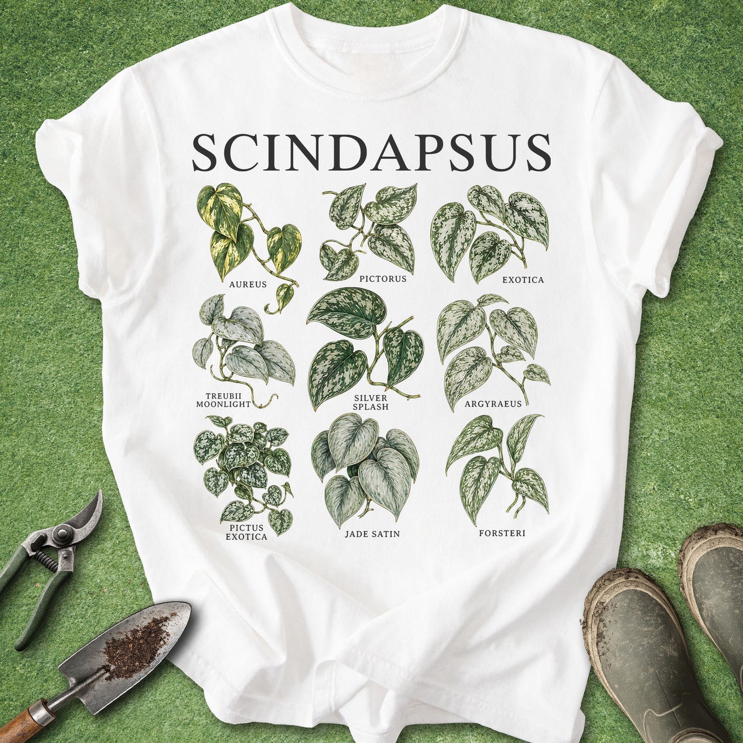 A white graphic T-shirt laid flat on green grass-like background, featuring the word 'SCINDAPSUS' and multiple illustrated leafy plant labels. Gardening props including pruning shears, a small hand trowel, and boots are placed around the shirt.