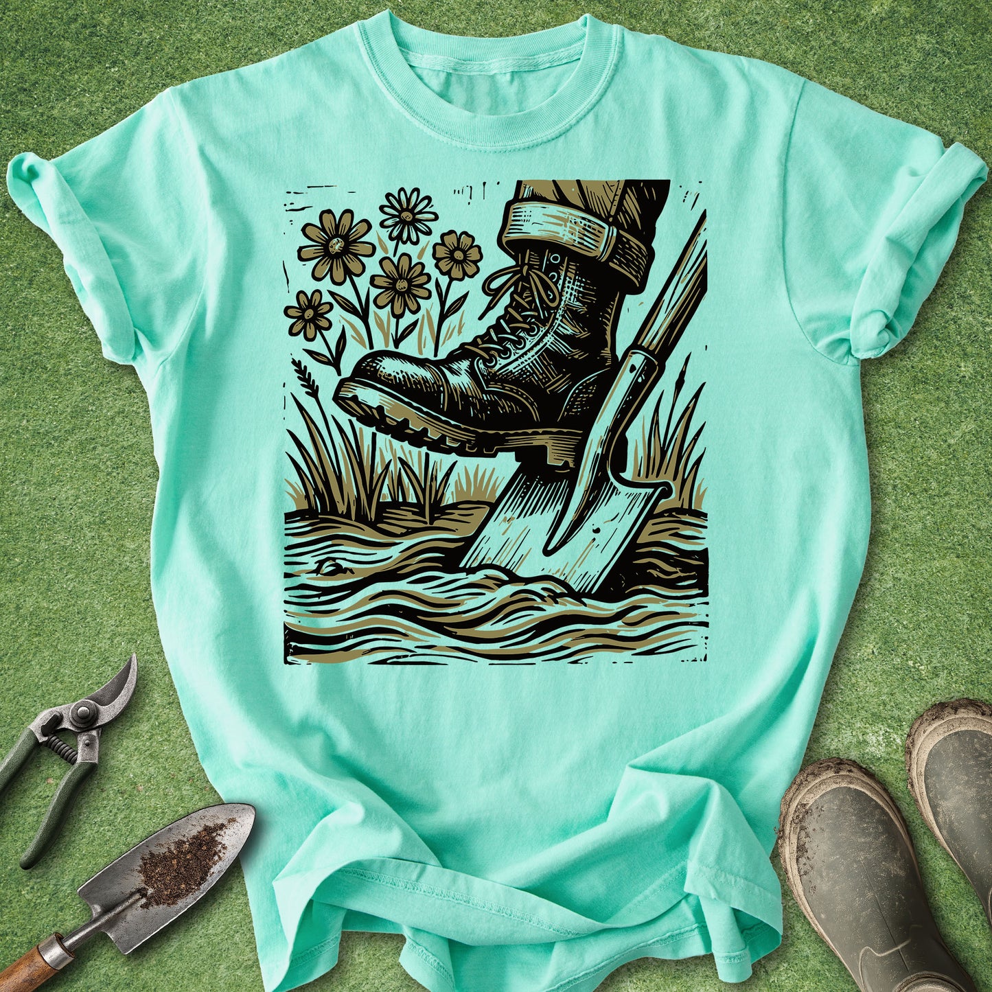 A mint green short-sleeve t-shirt laid flat on a green grass-like surface, featuring a large graphic print of a black work boot stepping into water with a shovel, surrounded by flowers and grass. Gardening tools and muddy boots are placed around the shirt as styled props.
