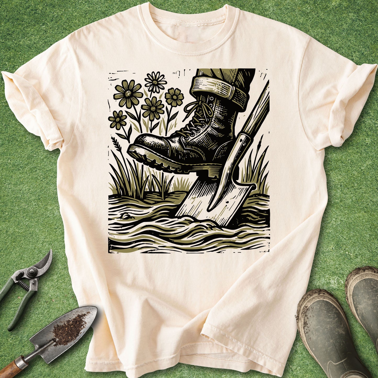 Comfort Colors ivory t-shirt featuring a vintage-style illustration of a gardening boot pressing on a shovel in the soil, surrounded by wildflowers and tall grass.
