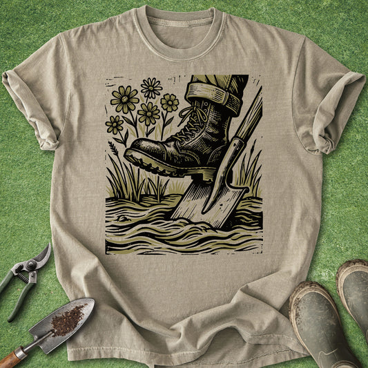 Comfort Colors khaki t-shirt featuring a vintage-style illustration of a gardening boot pressing on a shovel in the soil, surrounded by wildflowers and tall grass.