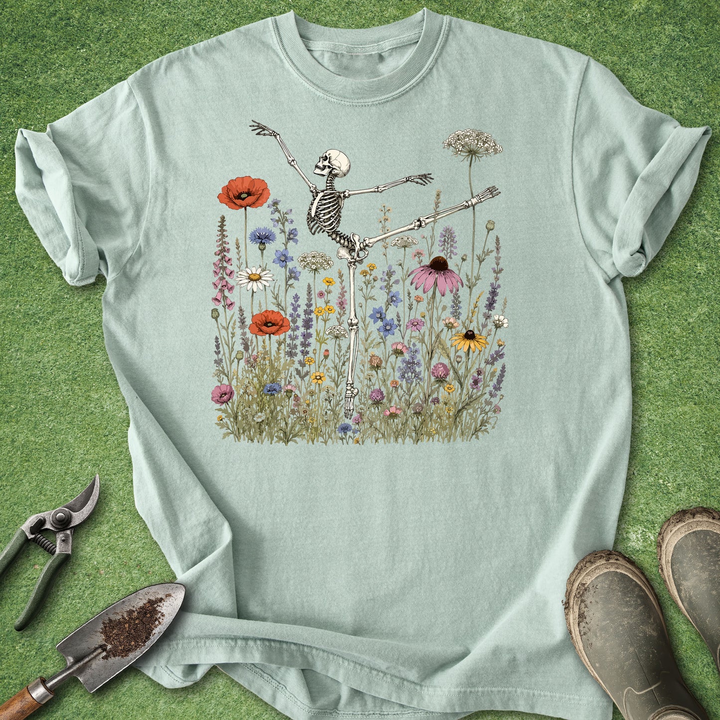 A light sage green short-sleeve t-shirt with a graphic of a skeleton dancing in a field of wildflowers, laid flat on a green grass-like surface with gardening tools and boots nearby.