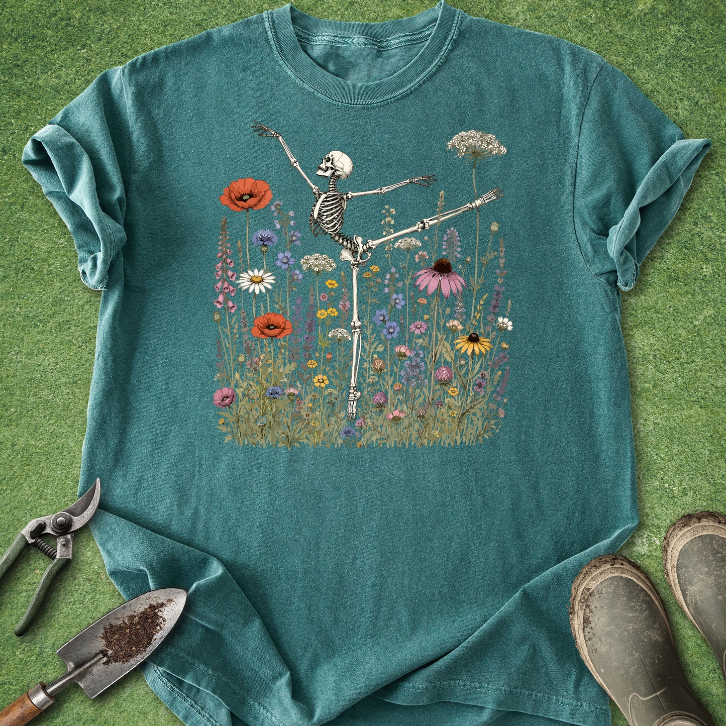 A teal short-sleeve t-shirt laid flat on a green grass-like surface, featuring a central graphic of a dancing skeleton surrounded by colorful wildflowers. Gardening tools and boots are placed around the shirt as styling props.