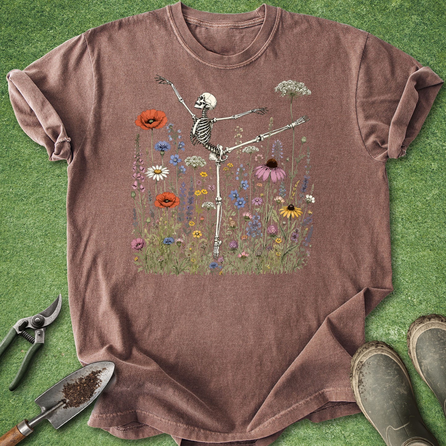 A mauve-brown short-sleeve t-shirt laid flat on green grass, featuring a central graphic of a dancing skeleton surrounded by colorful wildflowers. A gardening tool set and work boots are placed around the shirt as styling props.