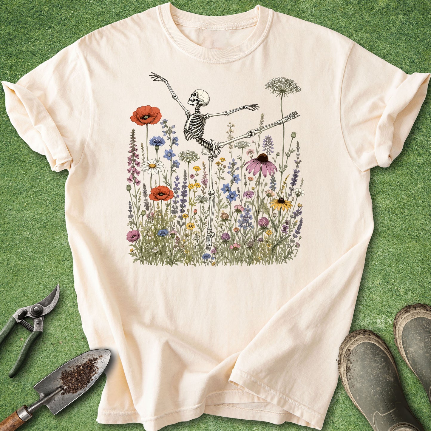 A cream-colored short-sleeve t-shirt with a graphic of a dancing skeleton surrounded by colorful wildflowers, laid flat on a green grass-like background with gardening tools and boots nearby.