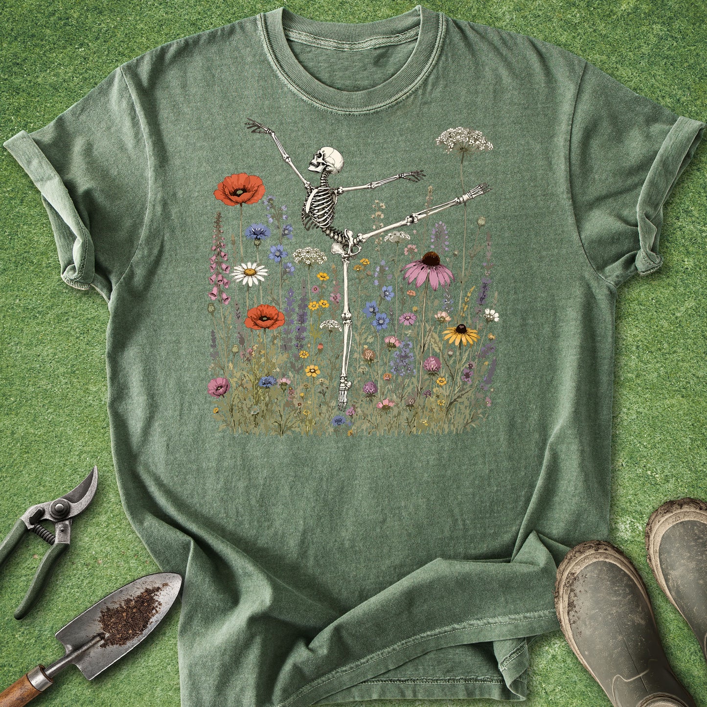 A sage green graphic t-shirt laid flat on a green grass-like surface, featuring a skeleton dancing among colorful wildflowers. Gardening tools and boots are styled around the shirt.