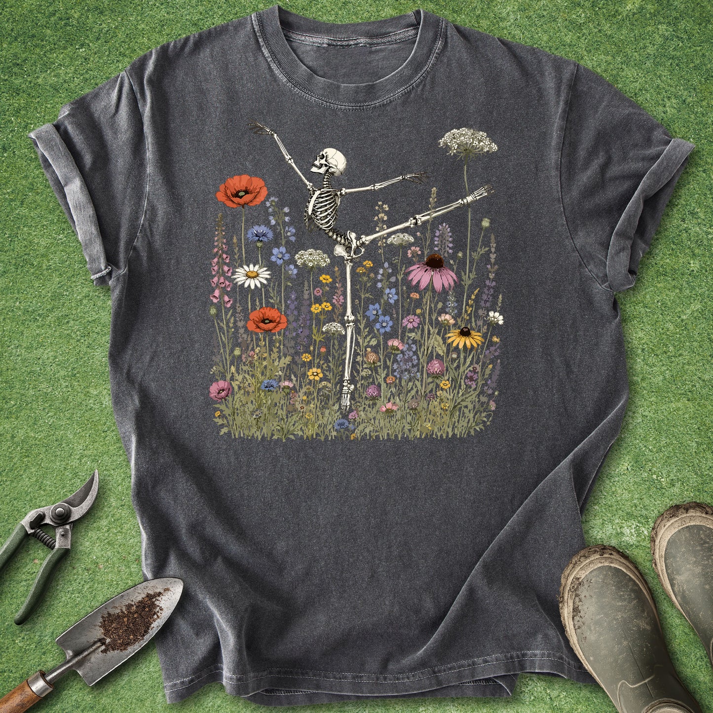 Pepper Comfort Colors t-shirt featuring an anatomically accurate skeleton performing a ballet arabesque pose surrounded by a colorful meadow of wildflowers including poppies, daisies, coneflowers, foxgloves, and Queen Anne's lace.