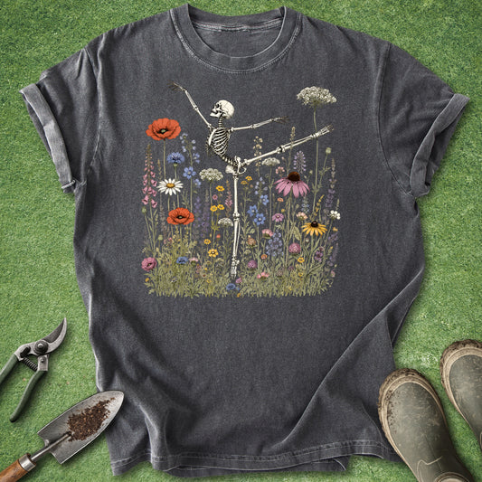 Pepper Comfort Colors t-shirt featuring an anatomically accurate skeleton performing a ballet arabesque pose surrounded by a colorful meadow of wildflowers including poppies, daisies, coneflowers, foxgloves, and Queen Anne's lace.