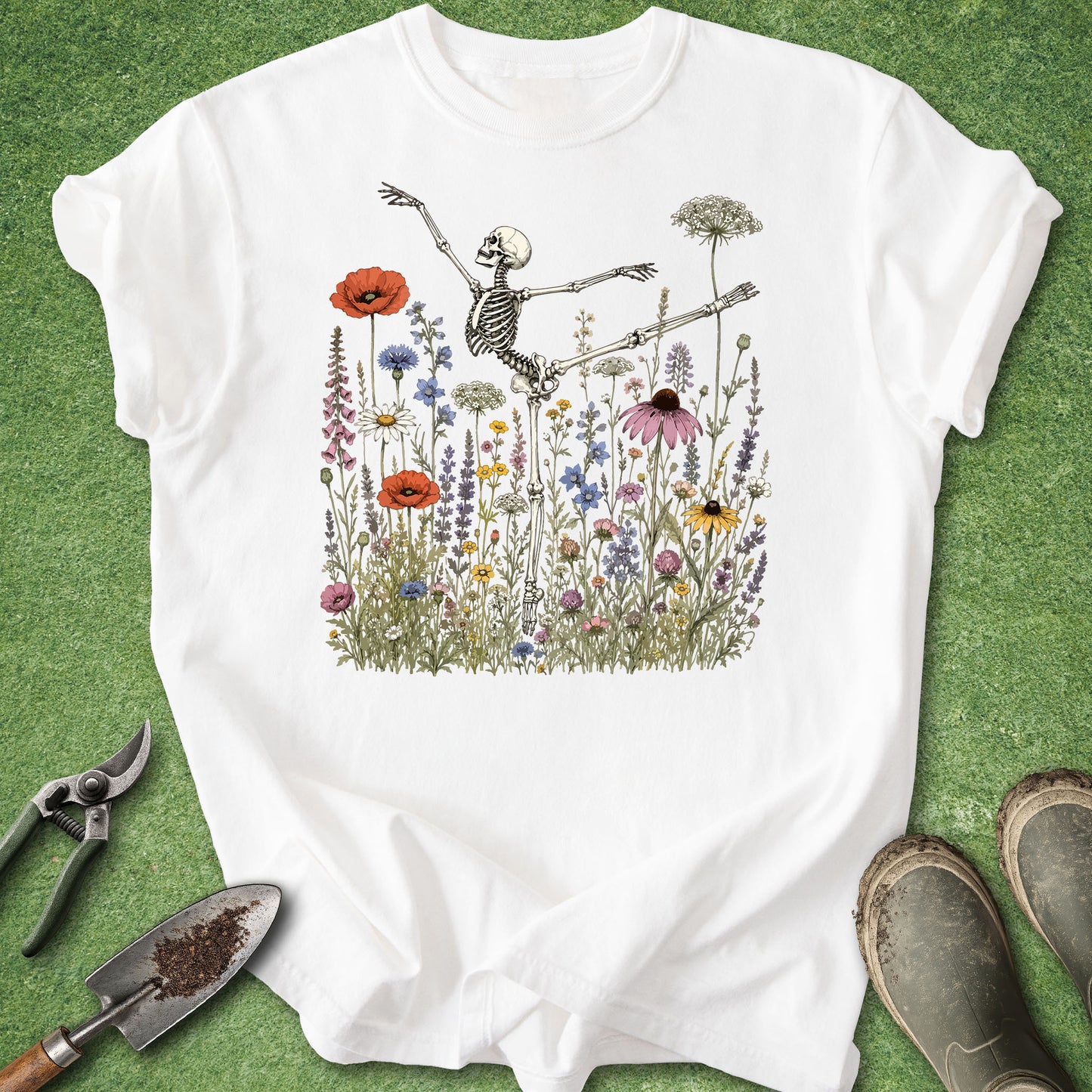 A white short-sleeve T-shirt laid flat on a green textured surface, featuring a central illustration of a skeleton dancing among colorful wildflowers. Gardening tools and muddy boots are placed around the shirt as styled props.