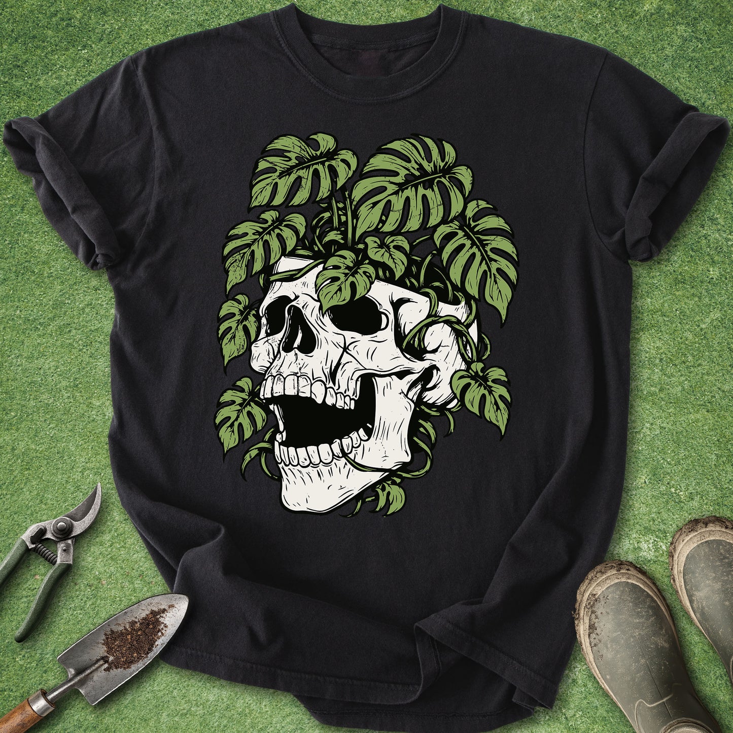 A black short-sleeve t-shirt laid flat on a green textured surface, featuring a graphic of a white skull surrounded by green monstera leaves. Gardening tools and boots are placed around the shirt as styling props.