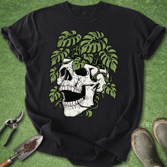A black short-sleeve t-shirt laid flat on a green textured surface, featuring a graphic of a white skull surrounded by green monstera leaves. Gardening tools and boots are placed around the shirt as styling props.