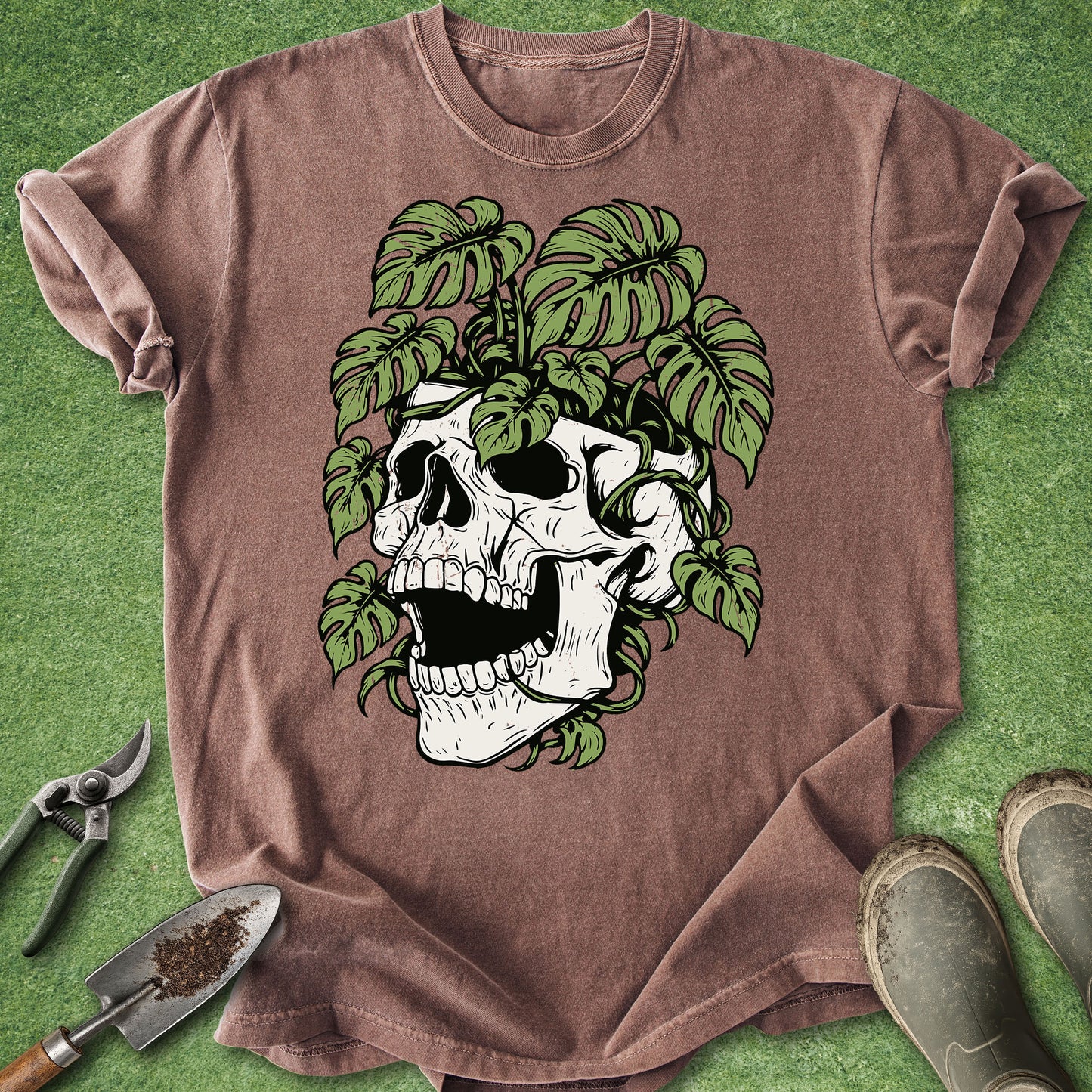 A brown short-sleeve t-shirt laid flat on green grass-like background, featuring a large graphic of a white skull surrounded by green monstera leaves. Gardening props including a trowel, pruning shears, and boots are visible around the shirt.