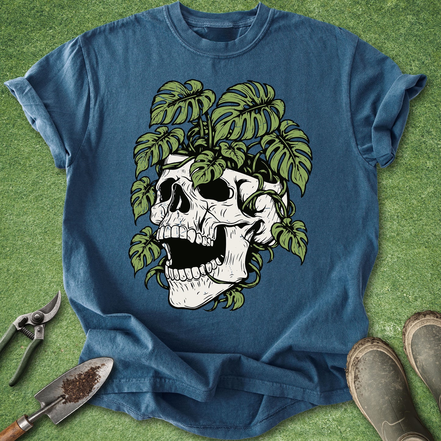 A blue short-sleeve t-shirt laid flat on green grass, featuring a large graphic of a white skull surrounded by green monstera leaves. Gardening tools and boots are placed around the shirt as styling props.