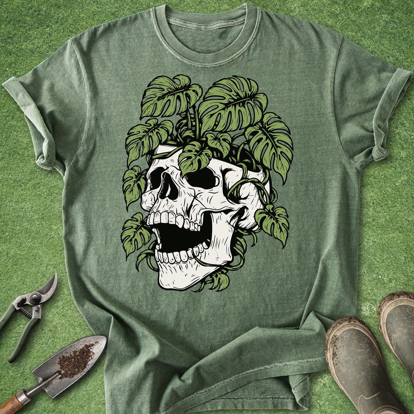 An olive green graphic t-shirt laid flat on a green grass-like surface, featuring a large white skull with green leaves growing out of it. Gardening tools and boots are styled around the shirt.