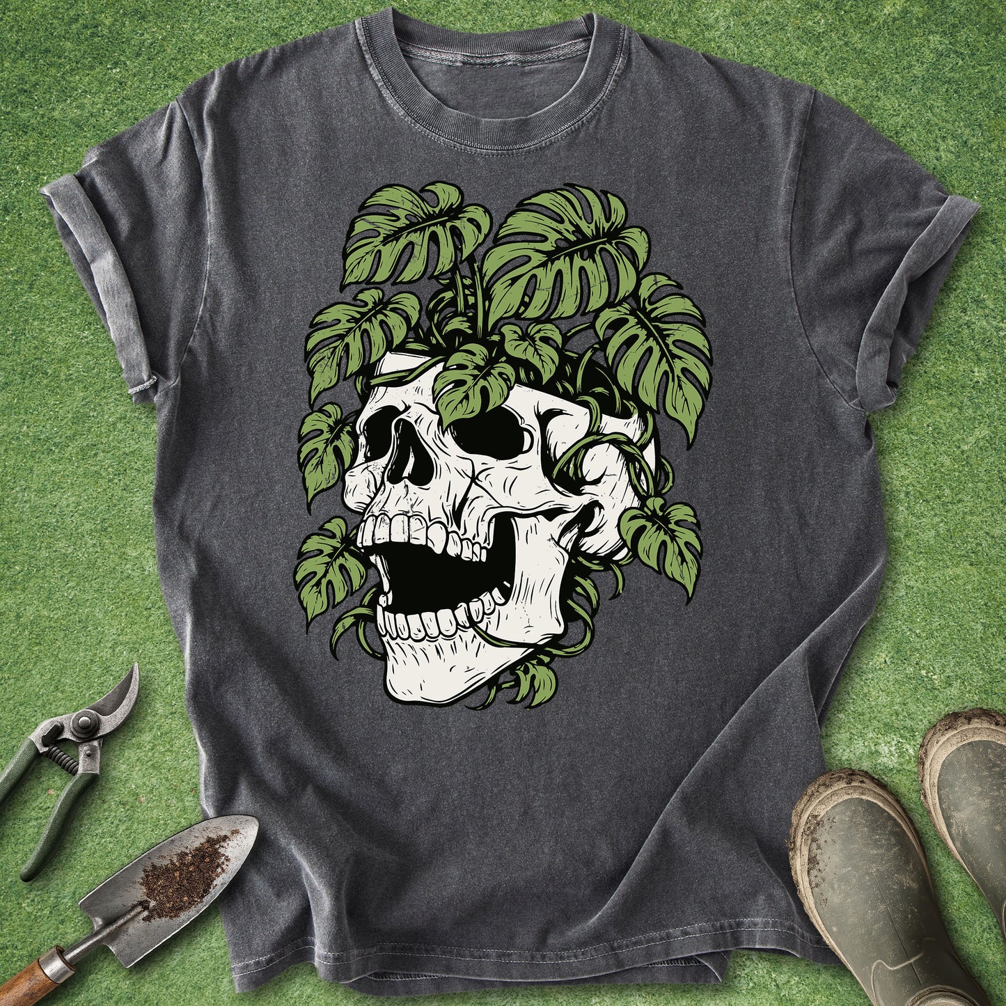 A dark gray graphic t-shirt featuring a white skull surrounded by green monstera leaves, laid flat on a green grass-like surface with gardening tools and boots nearby.